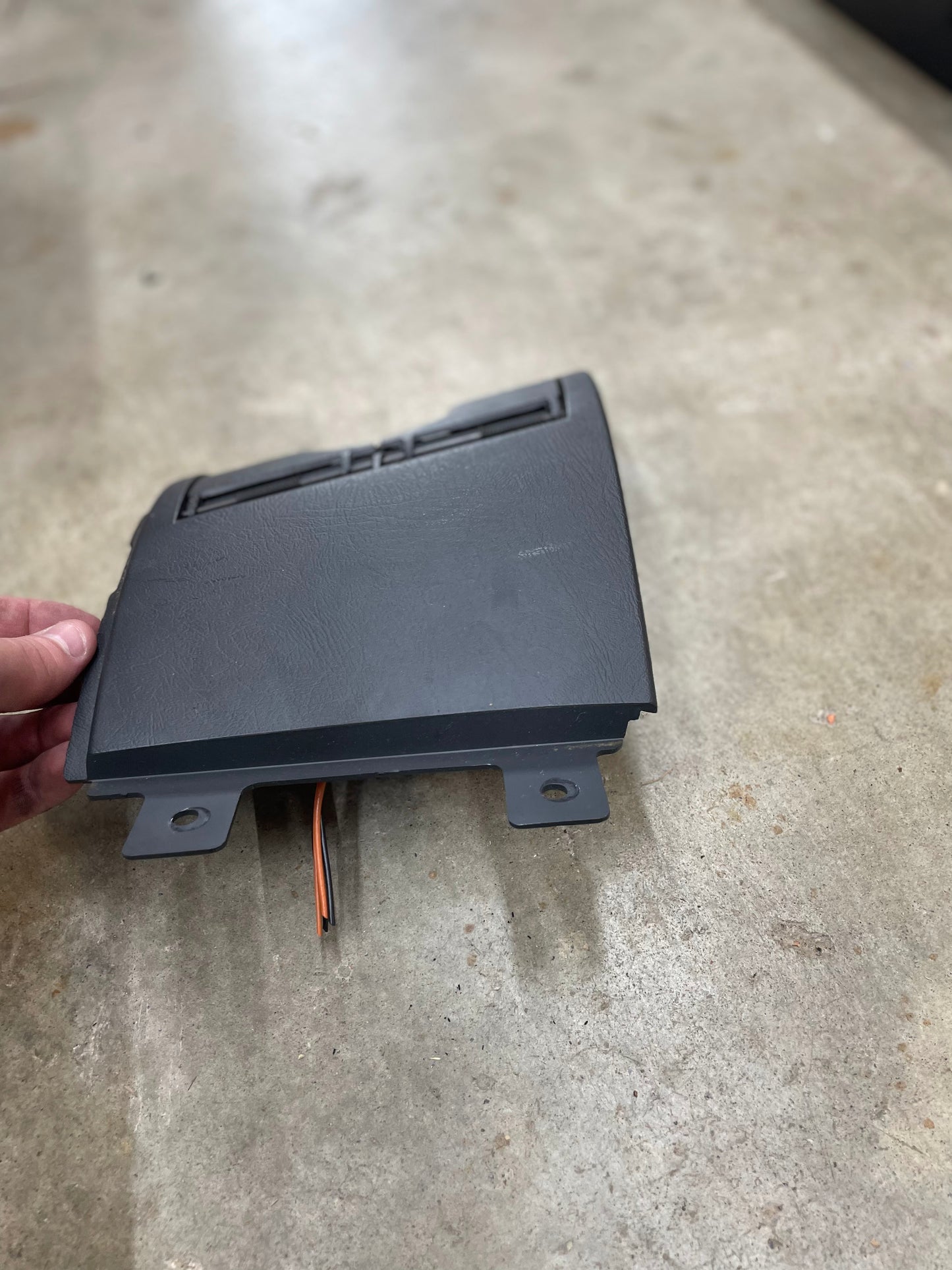 A hand holds a dark gray OEM 3 Port 12V Aux Panel Ash Tray Delete for a 1998-2000 GMC Envoy by General Motors, featuring two mounting tabs and wires, resting on a concrete floor.