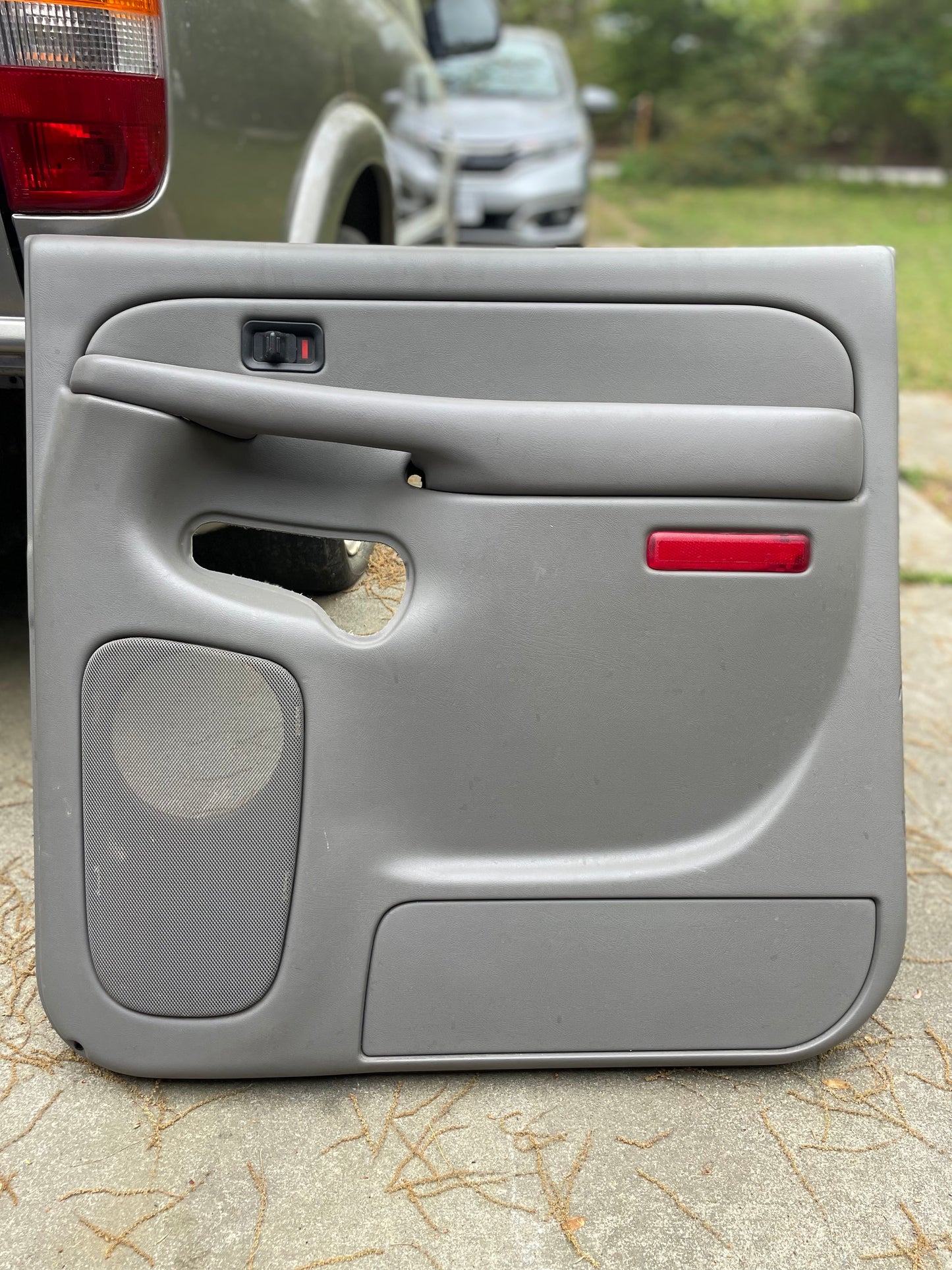 A General Motors OEM passenger side rear right leather door panel in light gray for 2003-2006 Chevy Suburban/Silverado Crew Cab is propped on concrete outdoors, featuring a speaker cutout, armrest, handle, and red reflector.