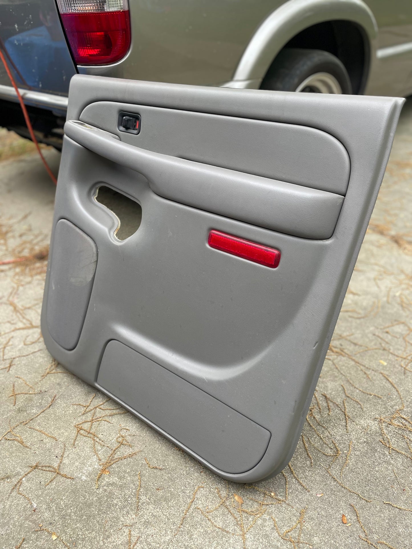 A General Motors OEM light gray leather passenger side rear right door panel for a 2003-2006 Chevy Suburban/Silverado Crew Cab sits on concrete outdoors; its handle is missing and a vehicle with a silver fender and red tail light is in the background.