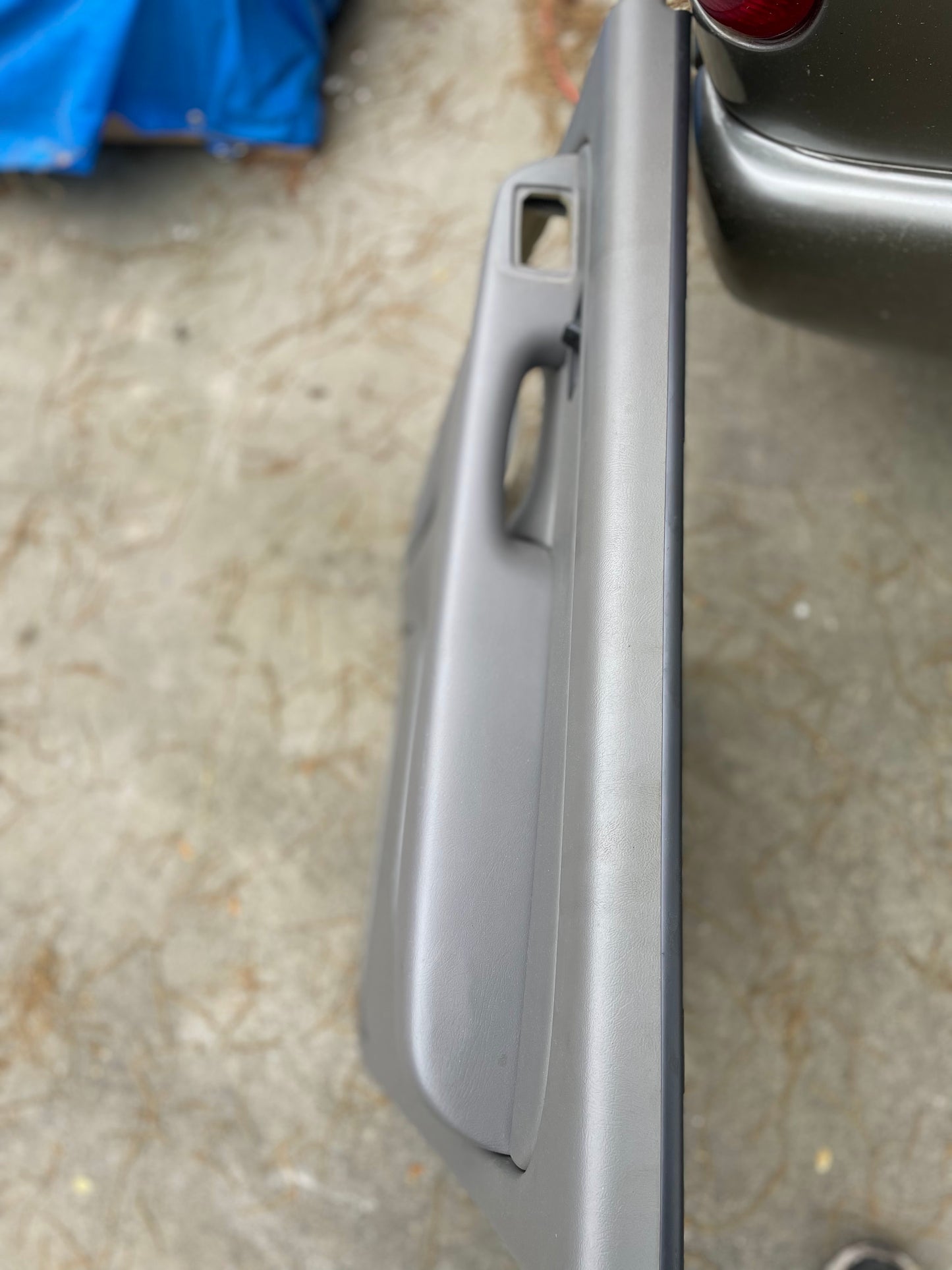A close-up of a General Motors OEM passenger side rear right-hand leather door panel in light gray, made for 2003-2006 Chevy Suburban and Silverado Crew Cab, is shown with handle detail; vehicle parked on concrete with pine needles nearby.