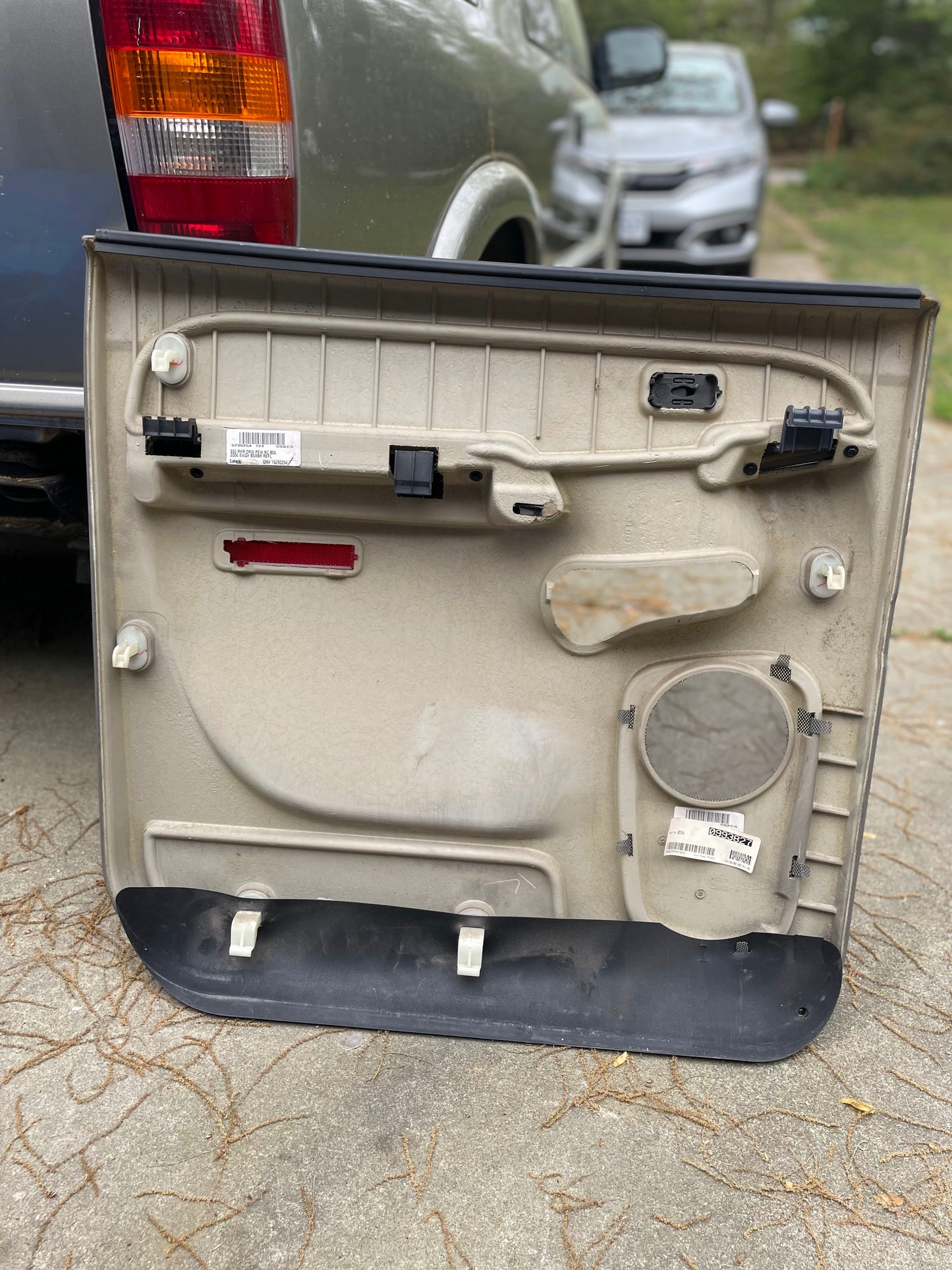 A General Motors OEM passenger side rear right hand leather door panel in light gray for 2003-2006 Chevy Suburban and Silverado Crew Cab lies on the ground by a parked vehicle, showing internal components and labels; another car is seen further ahead.