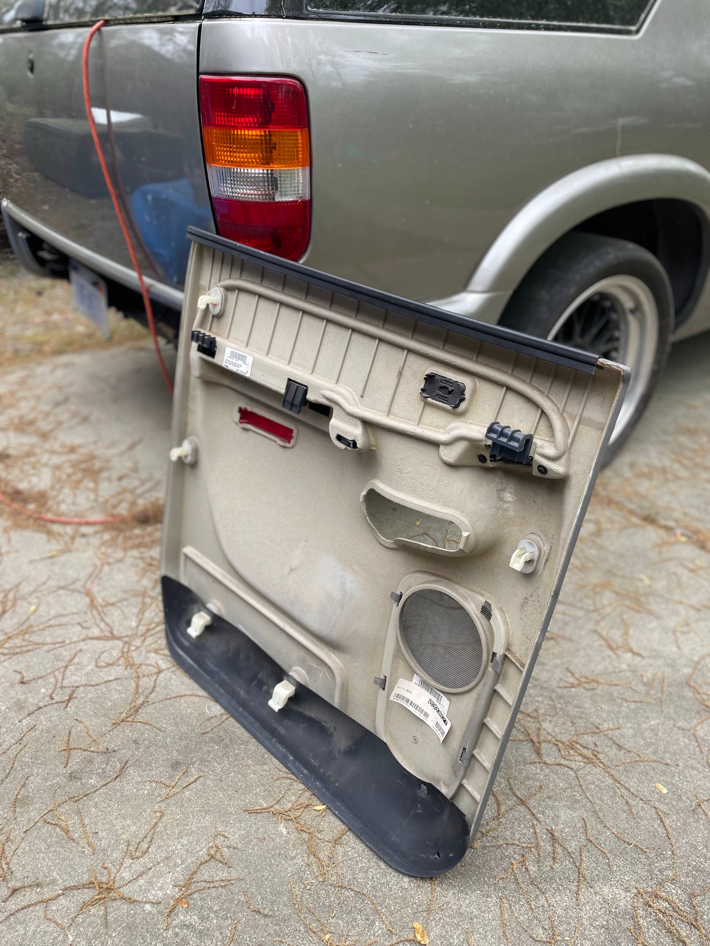 A General Motors OEM passenger side rear right hand leather door panel in light gray for 2003-2006 Chevy Suburban or Silverado Crew Cab rests near a gray SUV, showing wiring and clips, with the rear wheel and taillight visible behind.