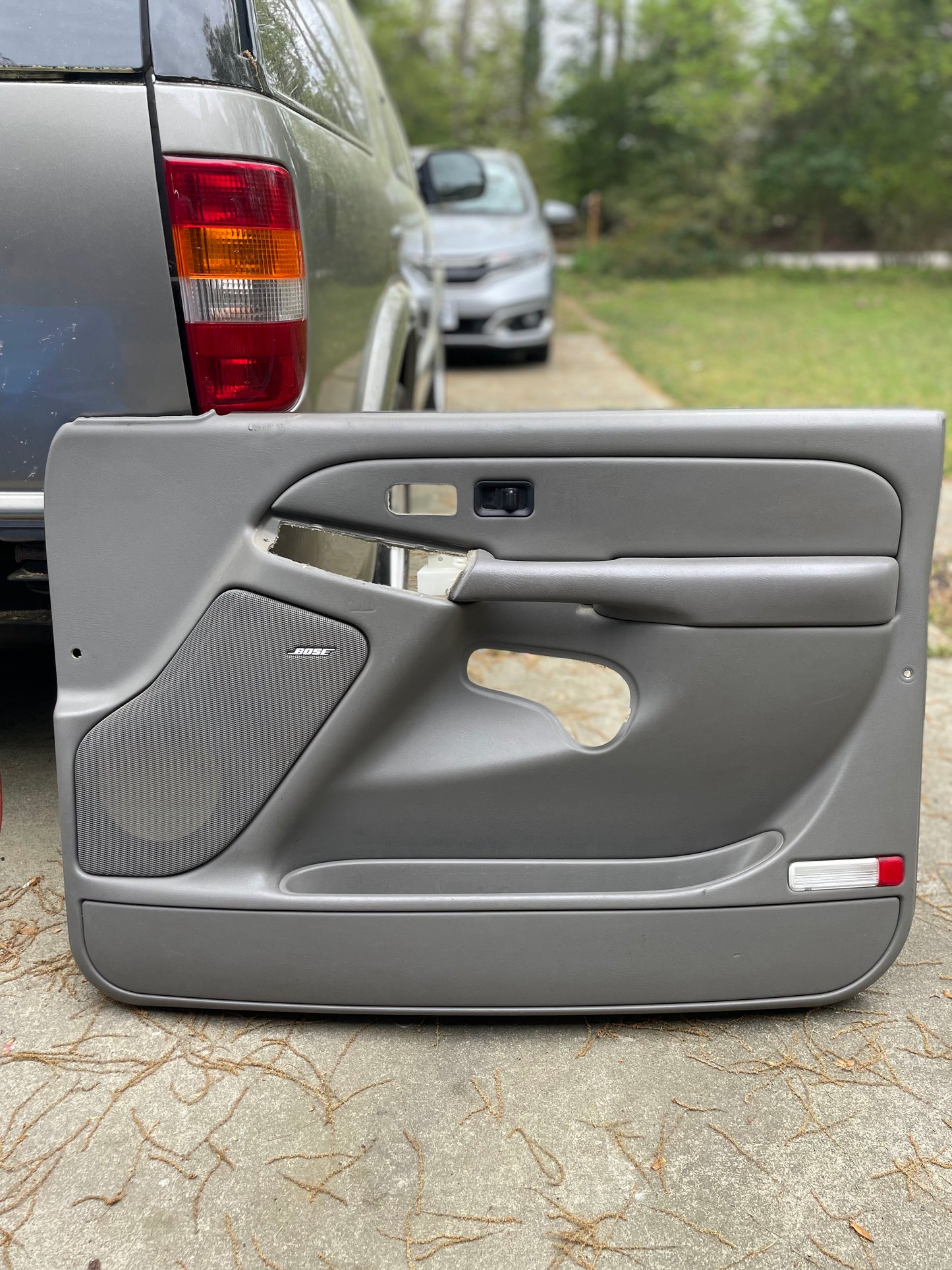A pair of OEM Light Gray Leather Door Trim Panels by General Motors for 2003-2006 Chevy Silverado/GMC Sierra rests on a driveway, with two vehicles—one gray, one silver—nearby and trees in the background.