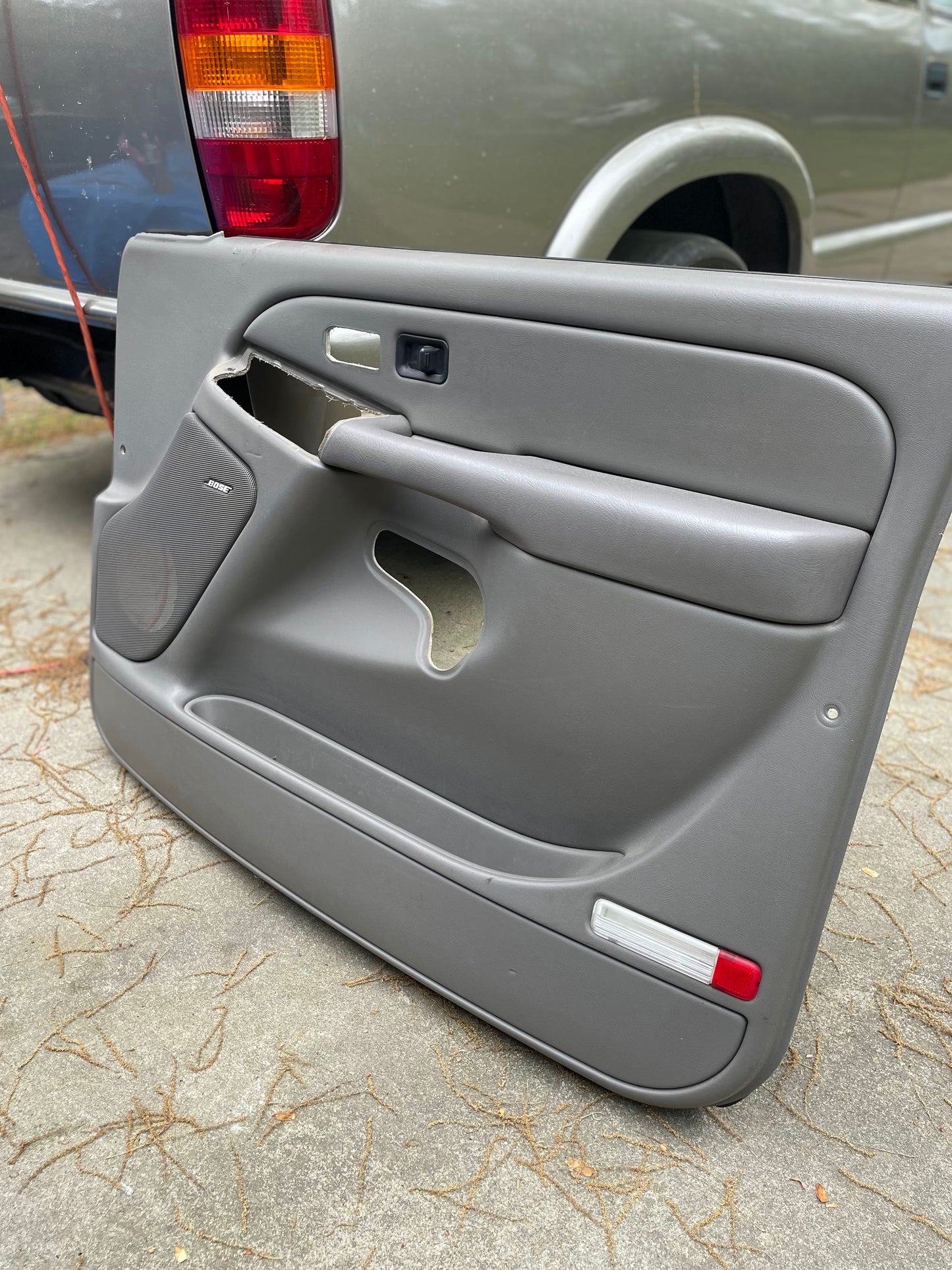 An OEM pair of General Motors front leather door trim panels in light gray for 2003-2006 Chevy Silverado/GMC Sierra is shown on the ground near a silver pickup, wiring visible against the pavement.