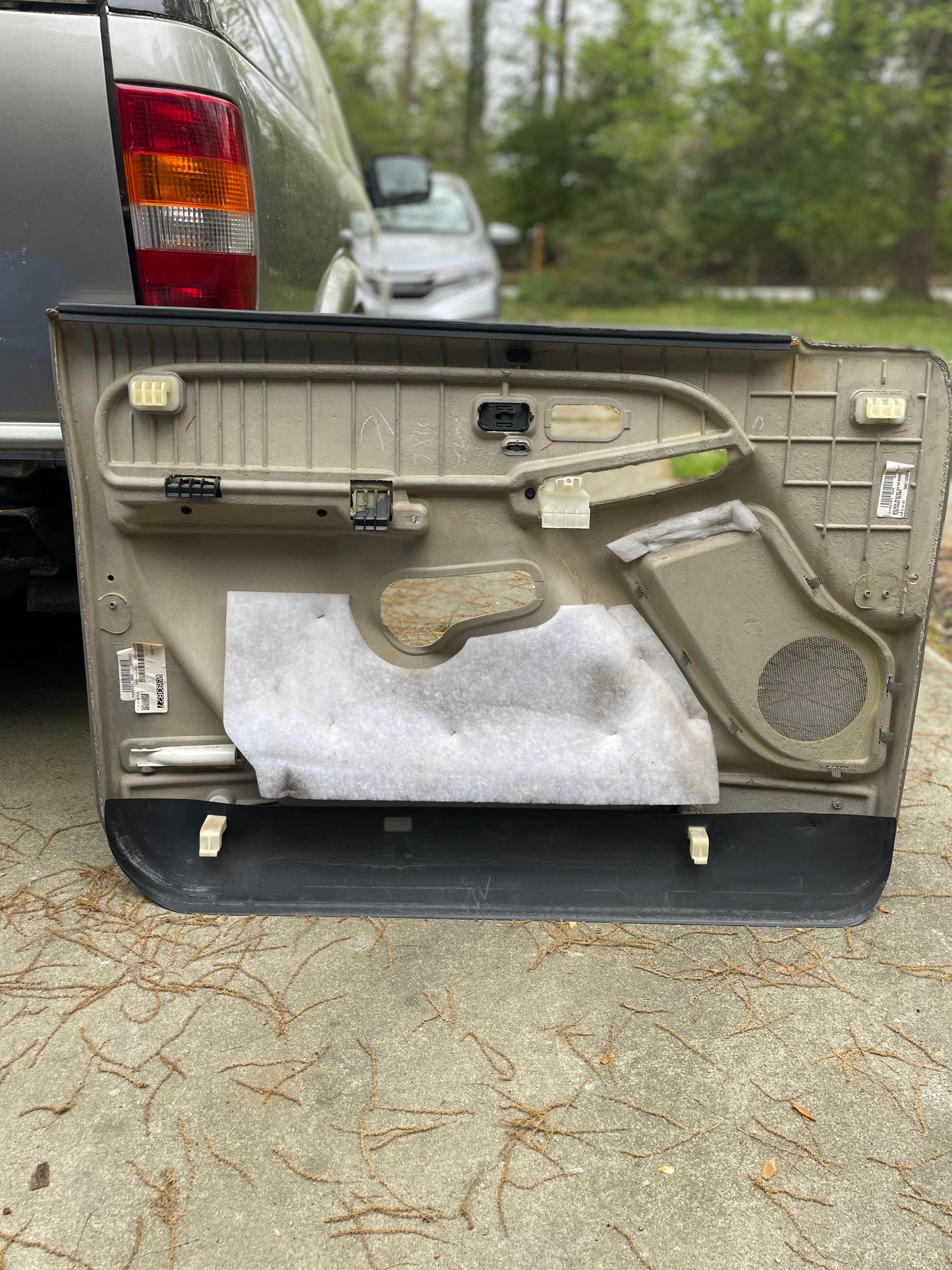 A pair of General Motors OEM front leather door trim panels in light gray for 2003-2006 Chevy Silverado and GMC Sierra, with inner mechanisms and wiring exposed, lies on the ground near a parked truck. Some insulation remains attached; trees are in the background.