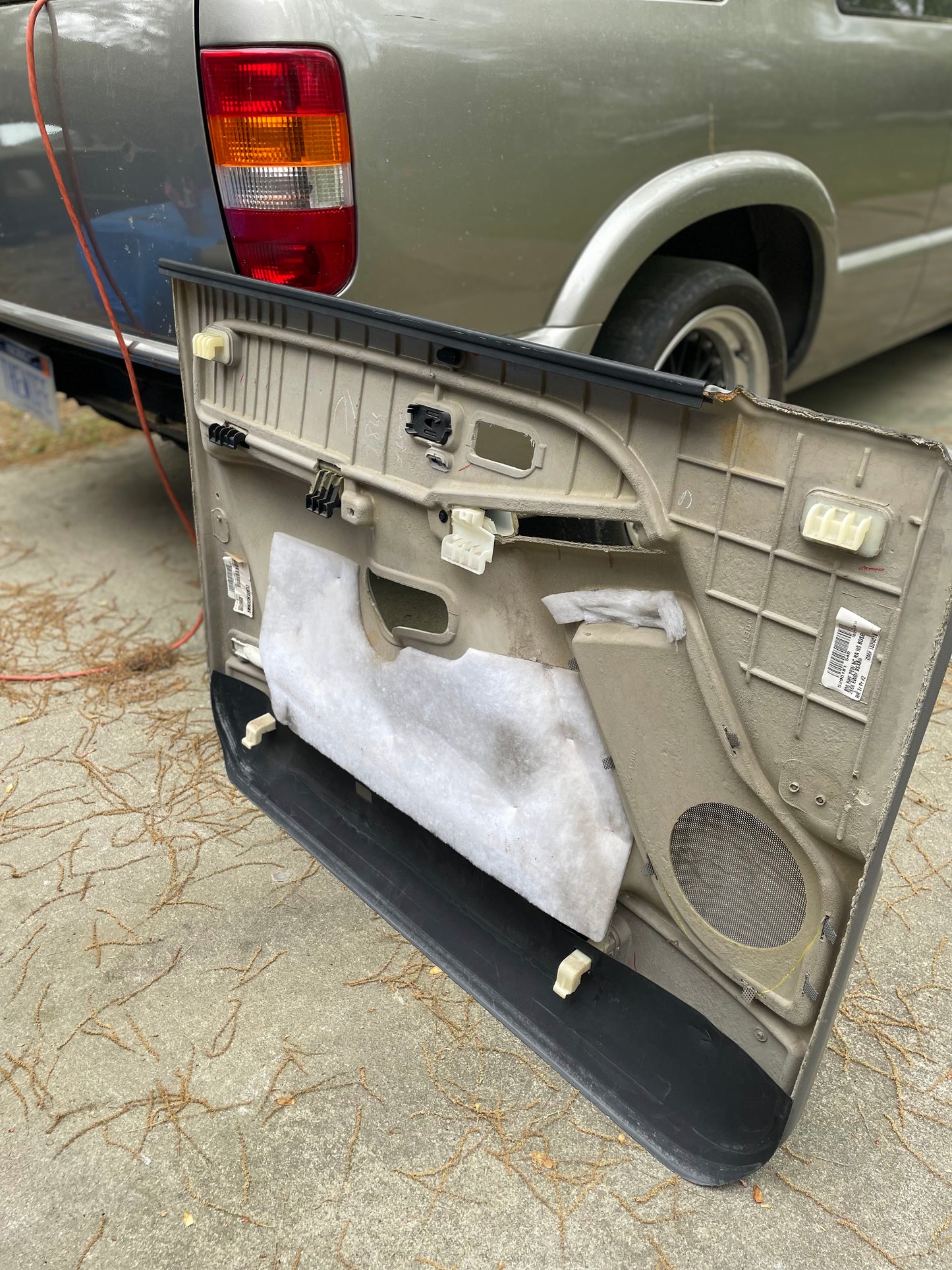 A pair of OEM Light Gray leather front door trim panels for 2003-2006 Chevy Silverado/GMC Sierra by General Motors rests against a silver GM truck in the driveway, exposing interior parts and insulation amid scattered pine needles.
