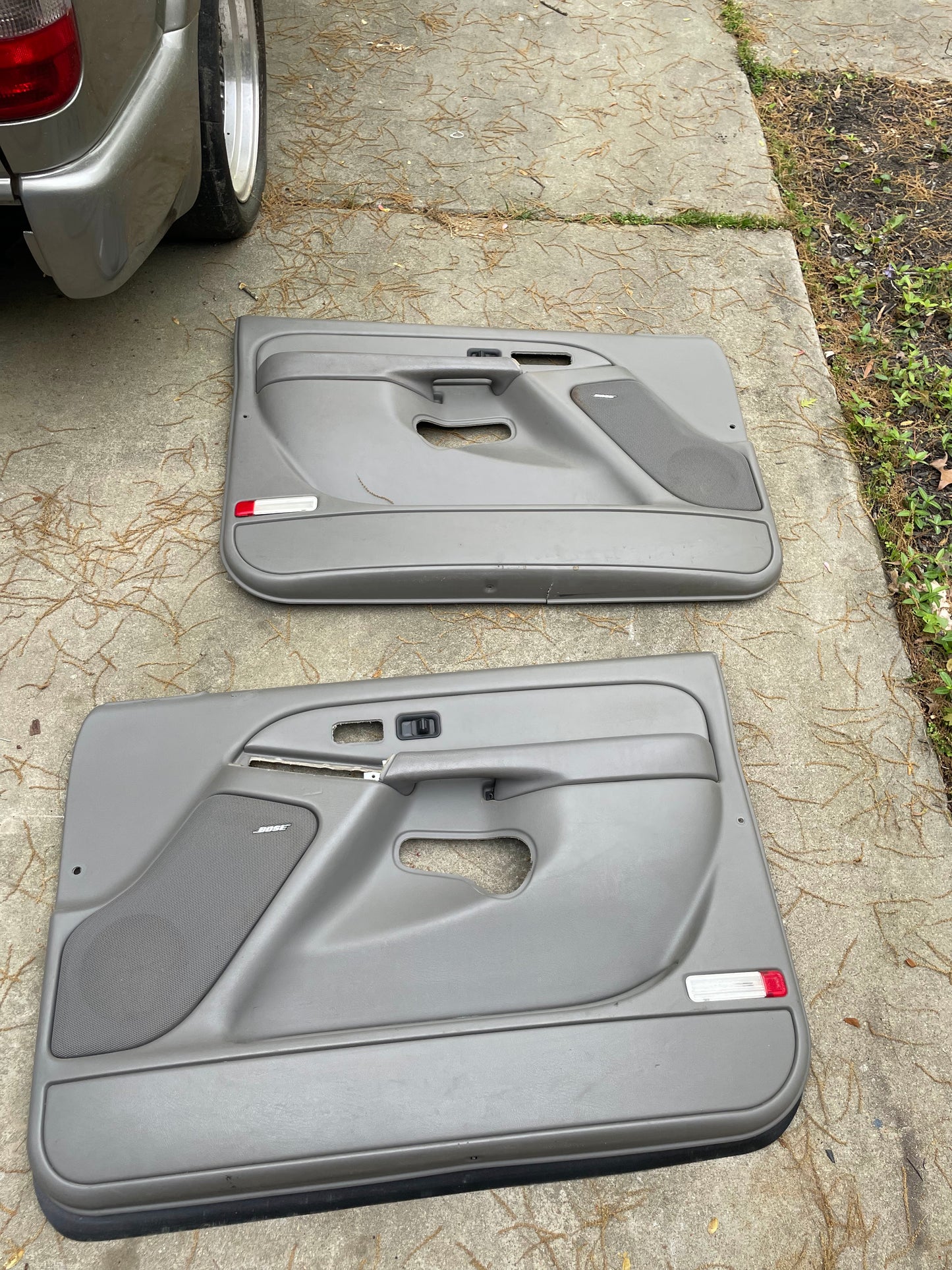 A pair of General Motors OEM front leather door trim panels in light gray, for 2003-2006 Chevy Silverado/GMC Sierra, are shown detached on a concrete driveway next to a silver vehicle.