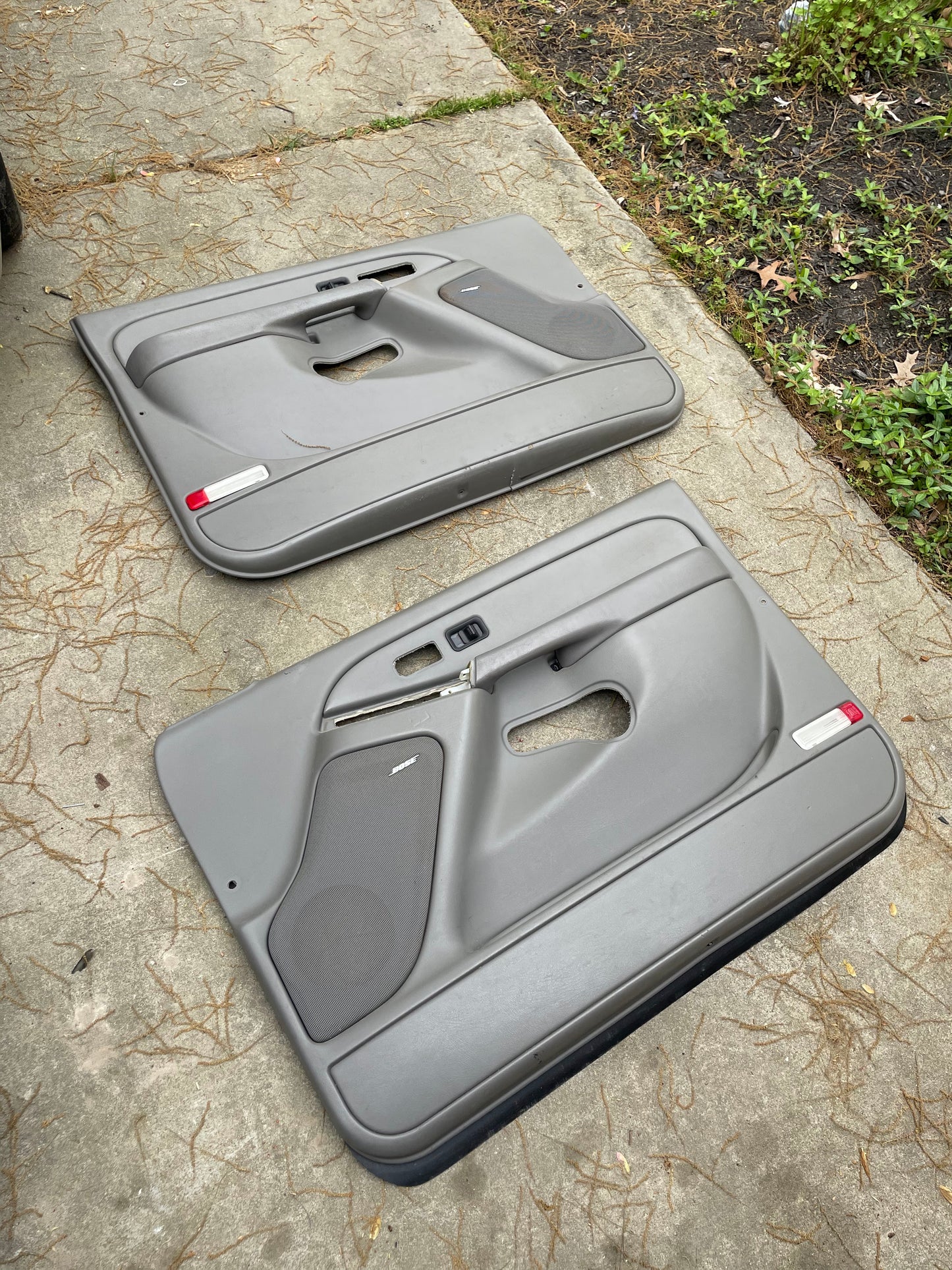 A pair of OEM General Motors front leather door trim panels in light gray for 2003-2006 Chevy Silverado and GMC Sierra, with speaker grilles and handle openings, are displayed outdoors on concrete beside soil and grass.