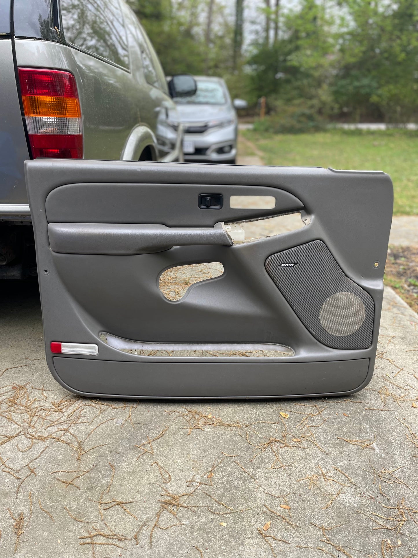 A pair of OEM General Motors front leather door trim panels in light gray, designed for 2003-2006 Chevy Silverado and GMC Sierra, rests upright outdoors on concrete; parked cars and trees are visible in the background.