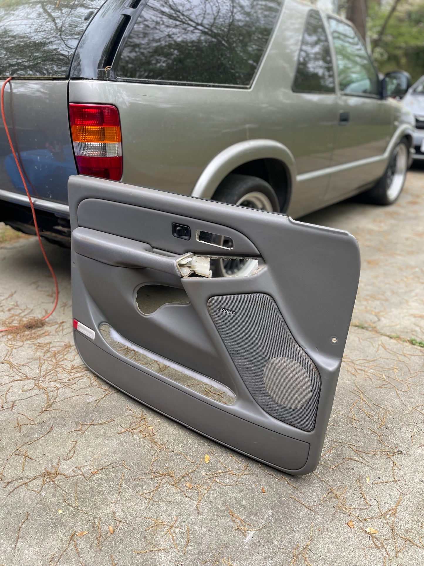 A pair of General Motors OEM front leather door trim panels in light gray, for 2003-2006 Chevy Silverado and GMC Sierra, sits next to a silver SUV with tree reflections in its windows. Some components and a speaker area are missing from the panels.