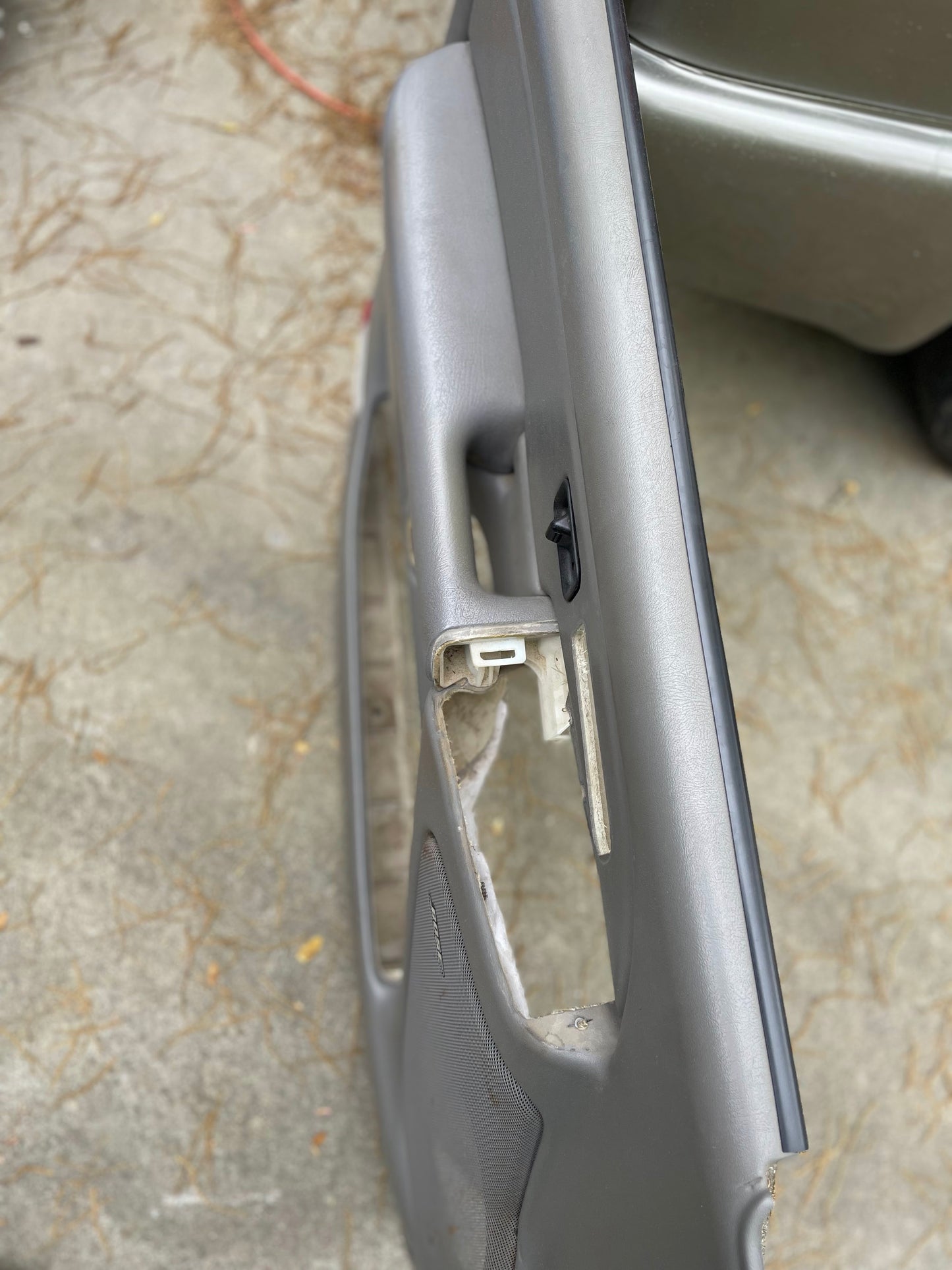 A close-up of an OEM pair of front leather door trim panels in light gray for 2003-2006 Chevy Silverado GMC Sierra by General Motors, showing missing handles and exposed components on a concrete surface scattered with pine needles.