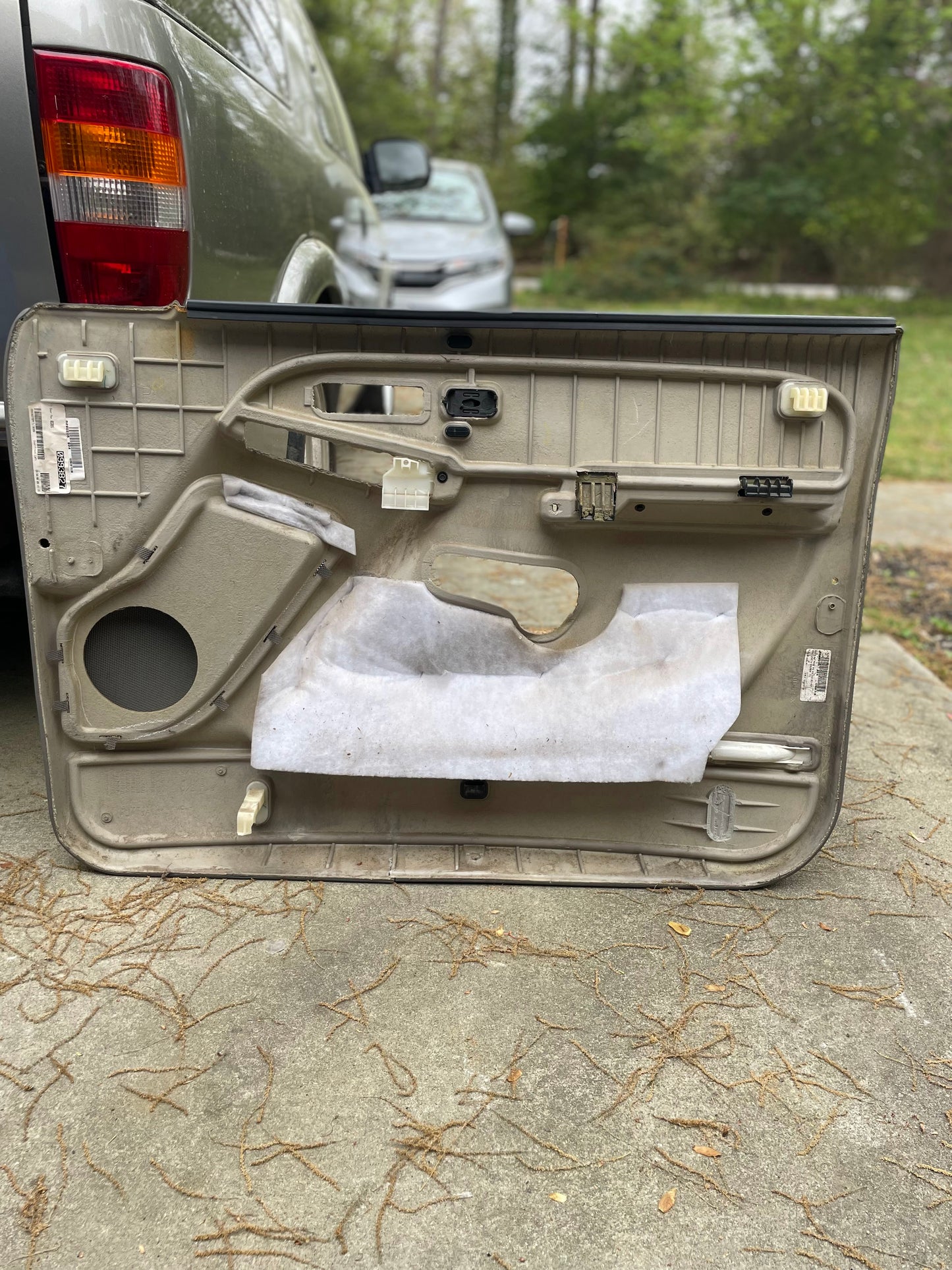 A pair of OEM General Motors front leather door trim panels in Light Gray for 2003-2006 Chevy Silverado and GMC Sierra is shown on the ground near a parked car, displaying the inner structure with another vehicle and greenery behind.