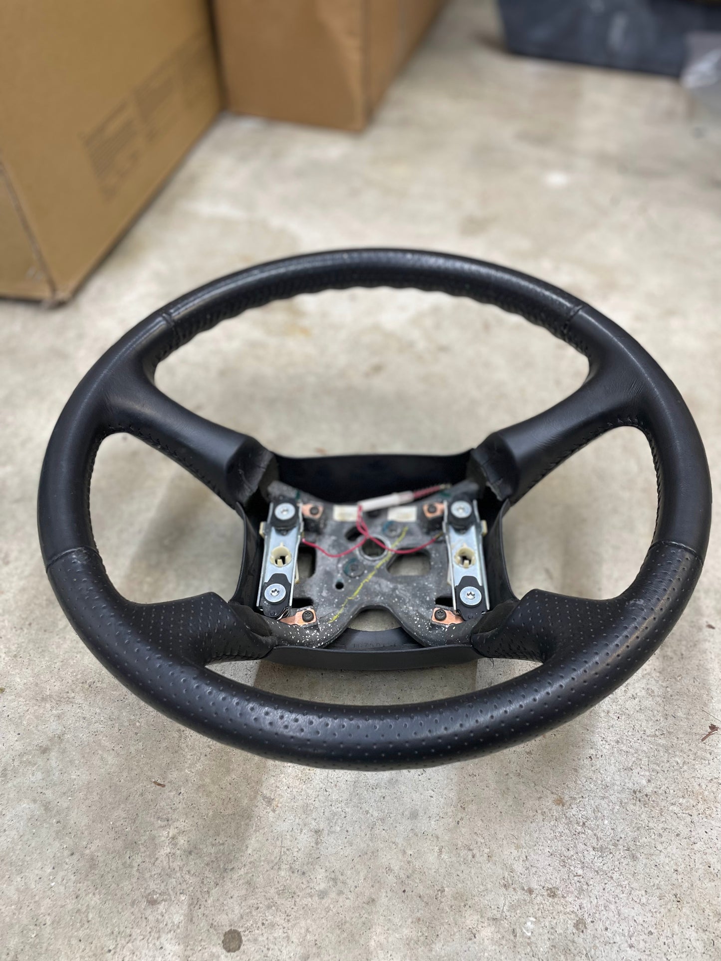 An OEM Perforated Leather Steering Wheel for 1999-2005 GM Chevy Silverado S10 by General Motors, missing its center cover and revealing wiring and mount points, sits on a concrete floor with cardboard boxes visible in the background.