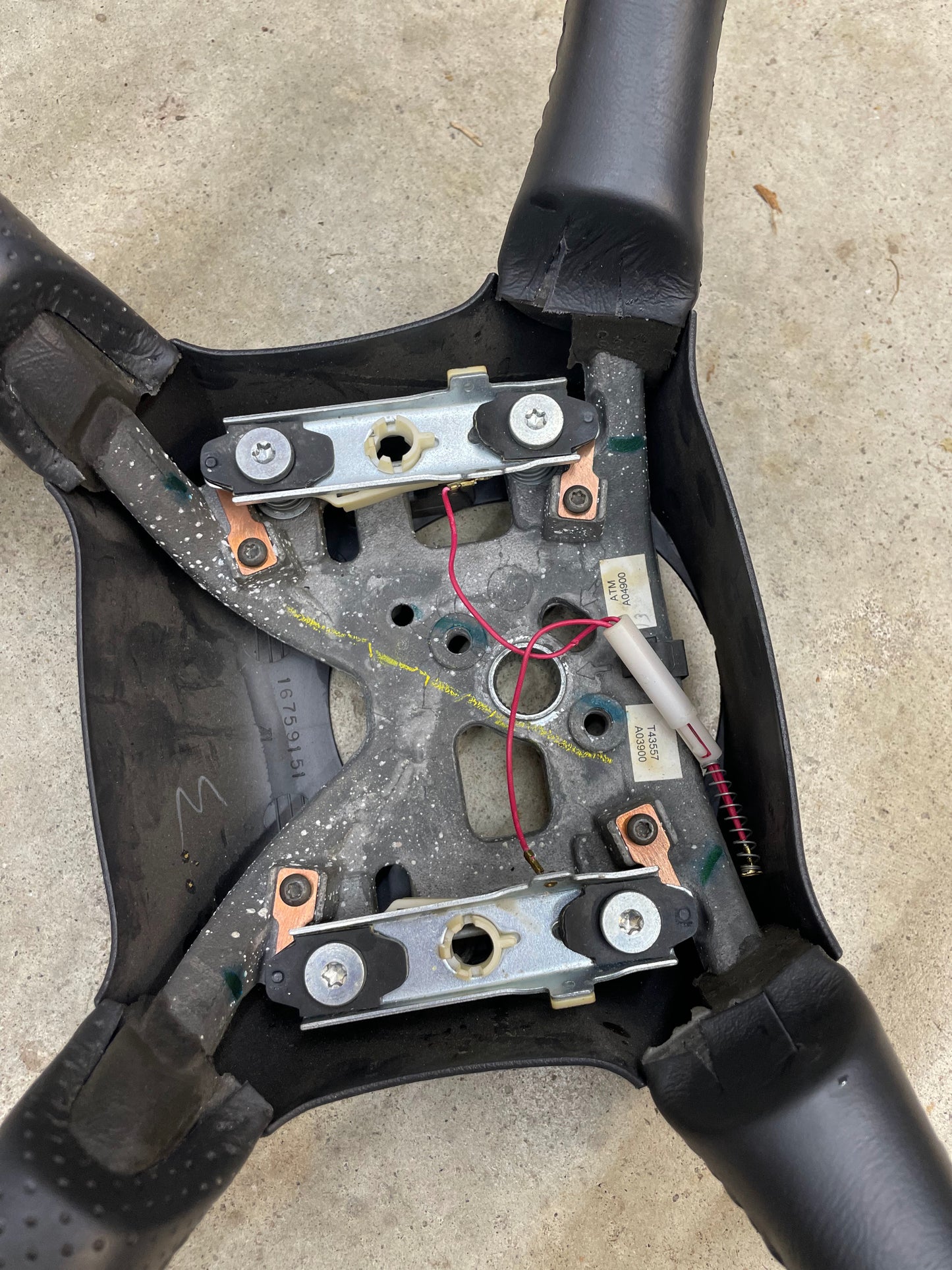 Close-up of the underside of an OEM Perforated Leather Steering Wheel by General Motors for 1999-2005 Chevy Silverado S10, showing exposed mechanics, wiring, metal brackets, and black grips on a concrete surface.