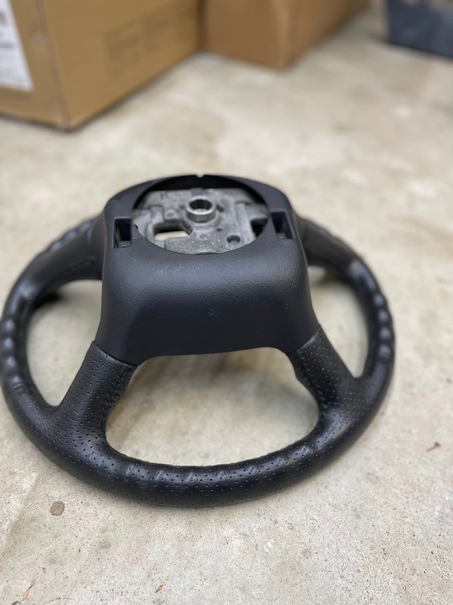 A General Motors OEM perforated leather steering wheel for 1999-2005 Chevy Silverado S10 and more, missing its center cover, rests on a concrete floor with cardboard boxes visible in the blurred background.