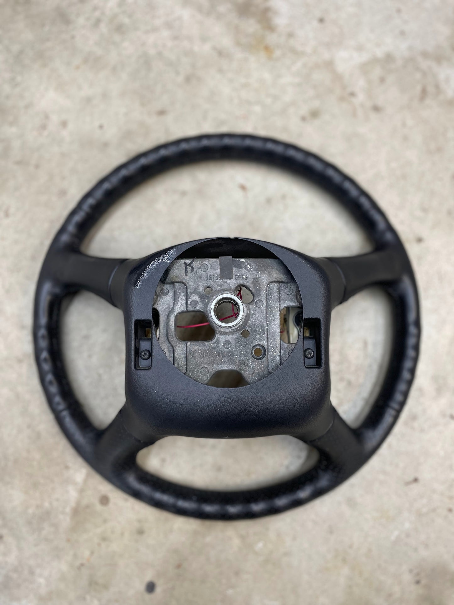 A General Motors OEM Perforated Leather Steering Wheel for 1999-2005 GM Chevy Silverado S10 is shown without its center, revealing metal components, placed on a light-colored concrete surface.