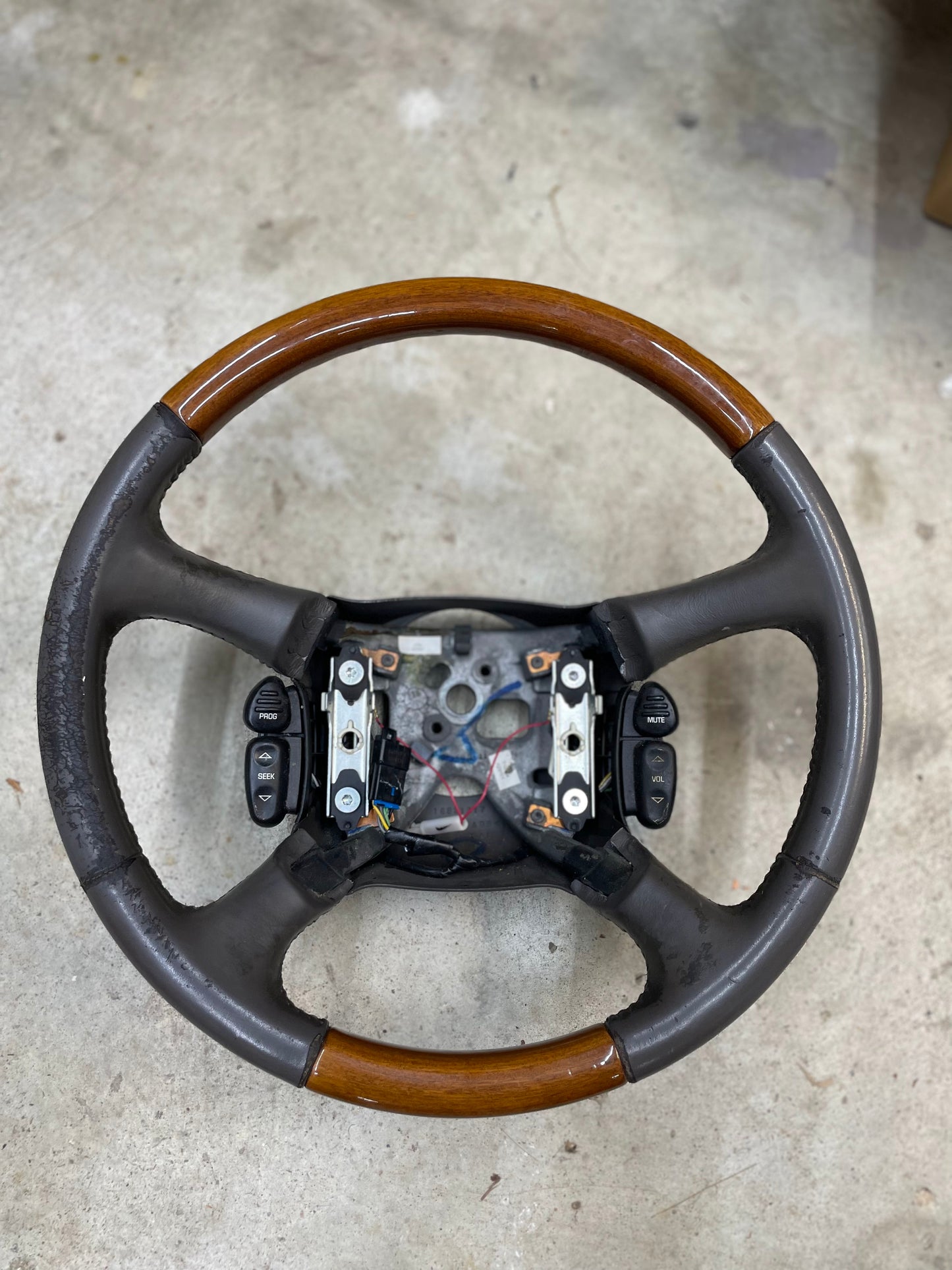 A General Motors OEM Leather w/Wood Steering Wheel with radio controls from an Escalade, compatible with 1995-2005 GM Trucks, rests on concrete missing its center horn pad and airbag, exposing wiring and internal mounts.