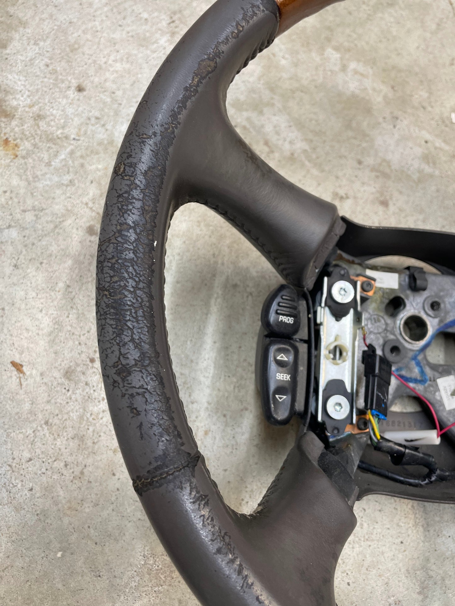 A close-up of a worn General Motors OEM Leather w/Wood Steering Wheel with radio controls from an Escalade (fits 1995-2005 GM Trucks), showing cracks, peeling, exposed wiring, and PROG/SEEK buttons.