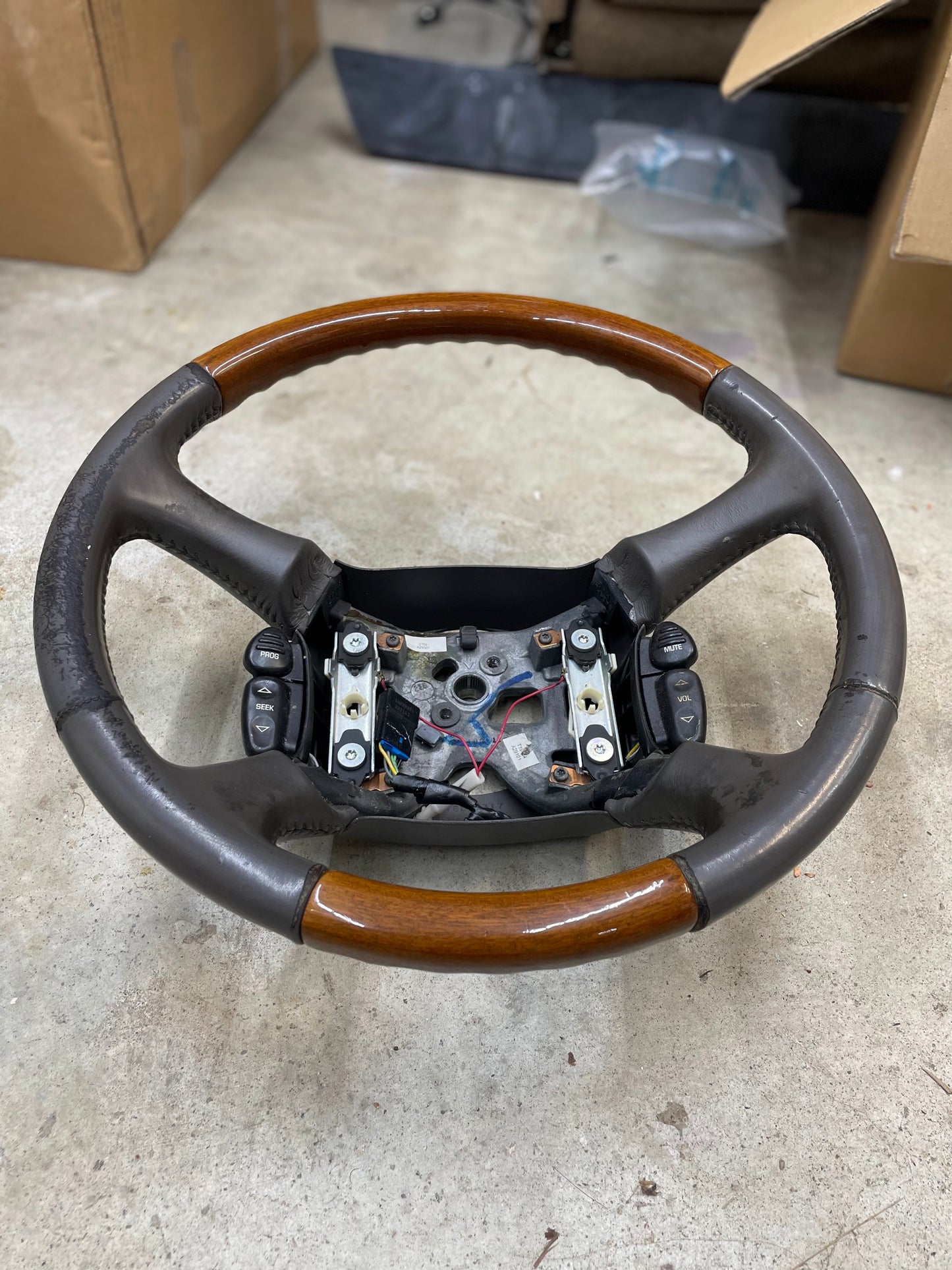 A General Motors OEM Leather w/Wood Steering Wheel with radio controls for 1995-2005 GM trucks, removed from an Escalade, rests on a concrete floor with exposed mounting points and wires; cardboard boxes and a plastic bag are visible behind it.