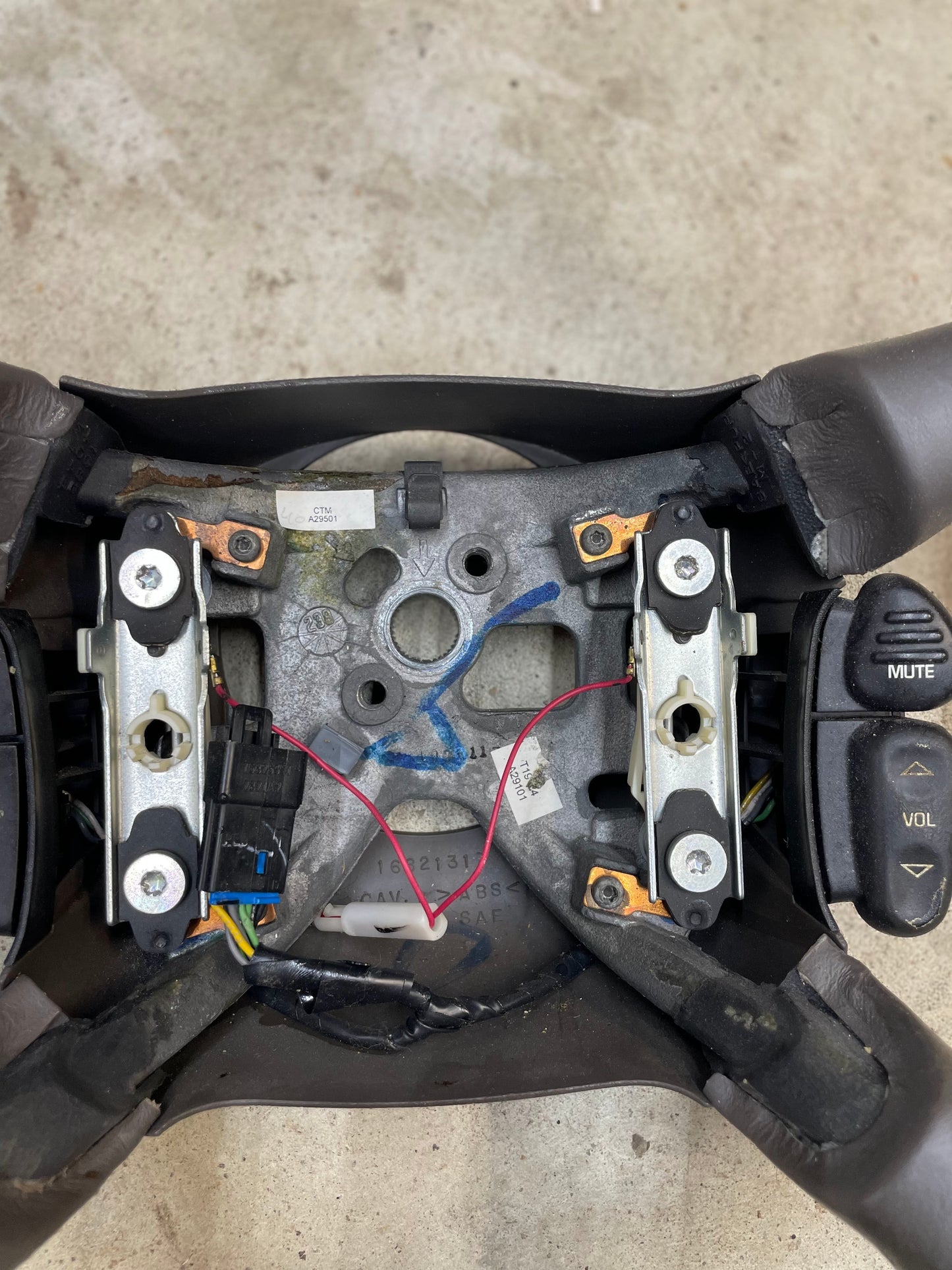 A close-up of a General Motors OEM Leather w/Wood Steering Wheel & Radio Controls from Escalade (fits 1995–2005 GM Trucks) shows the center cover removed, exposing internal parts and radio buttons on the right side.