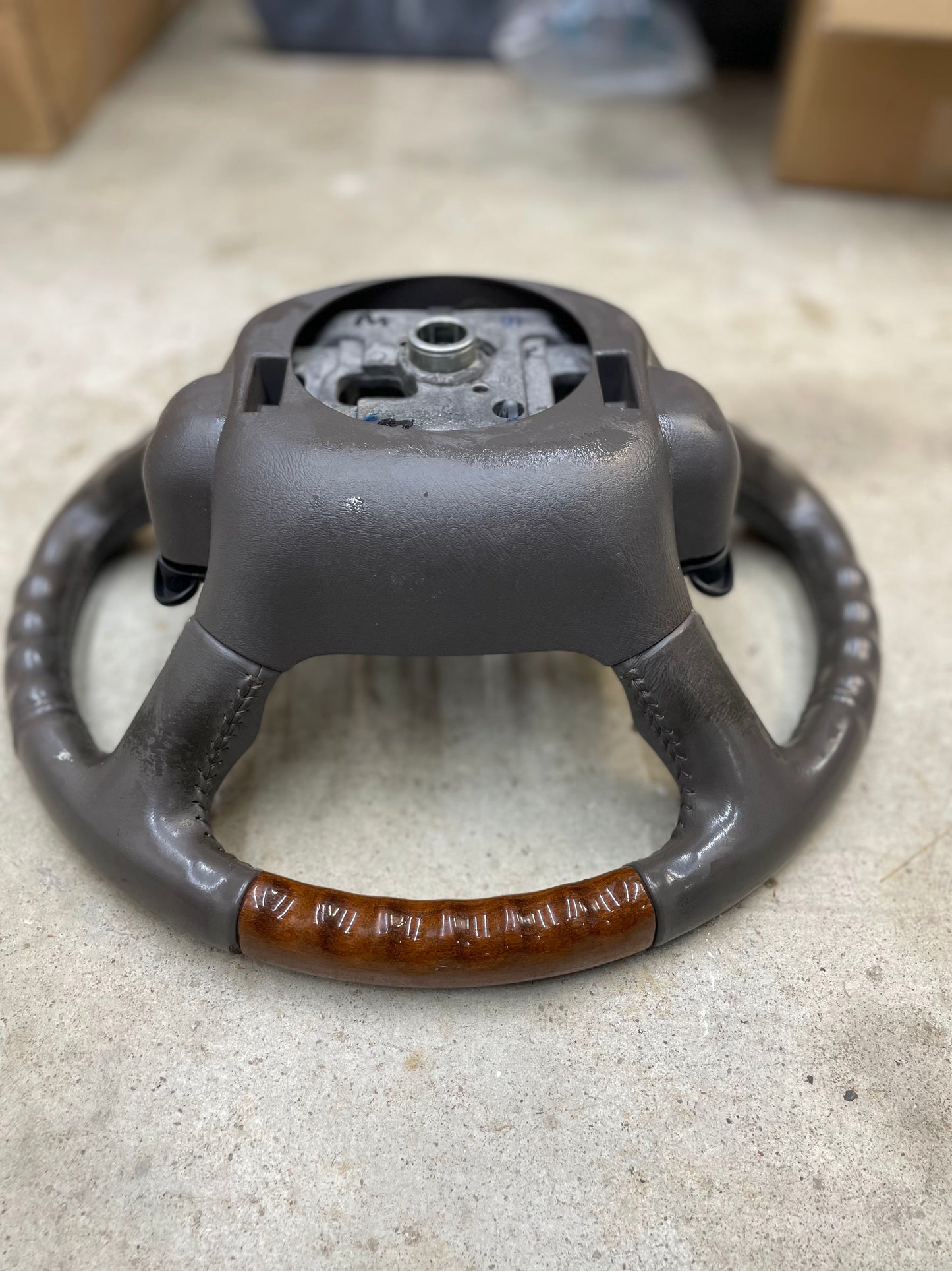 A General Motors OEM Leather w/Wood Steering Wheel with radio controls from an Escalade, fitting 1995–2005 GM Trucks, rests on a concrete floor with brown leather grip, wood bottom trim, and exposed mounting hardware.