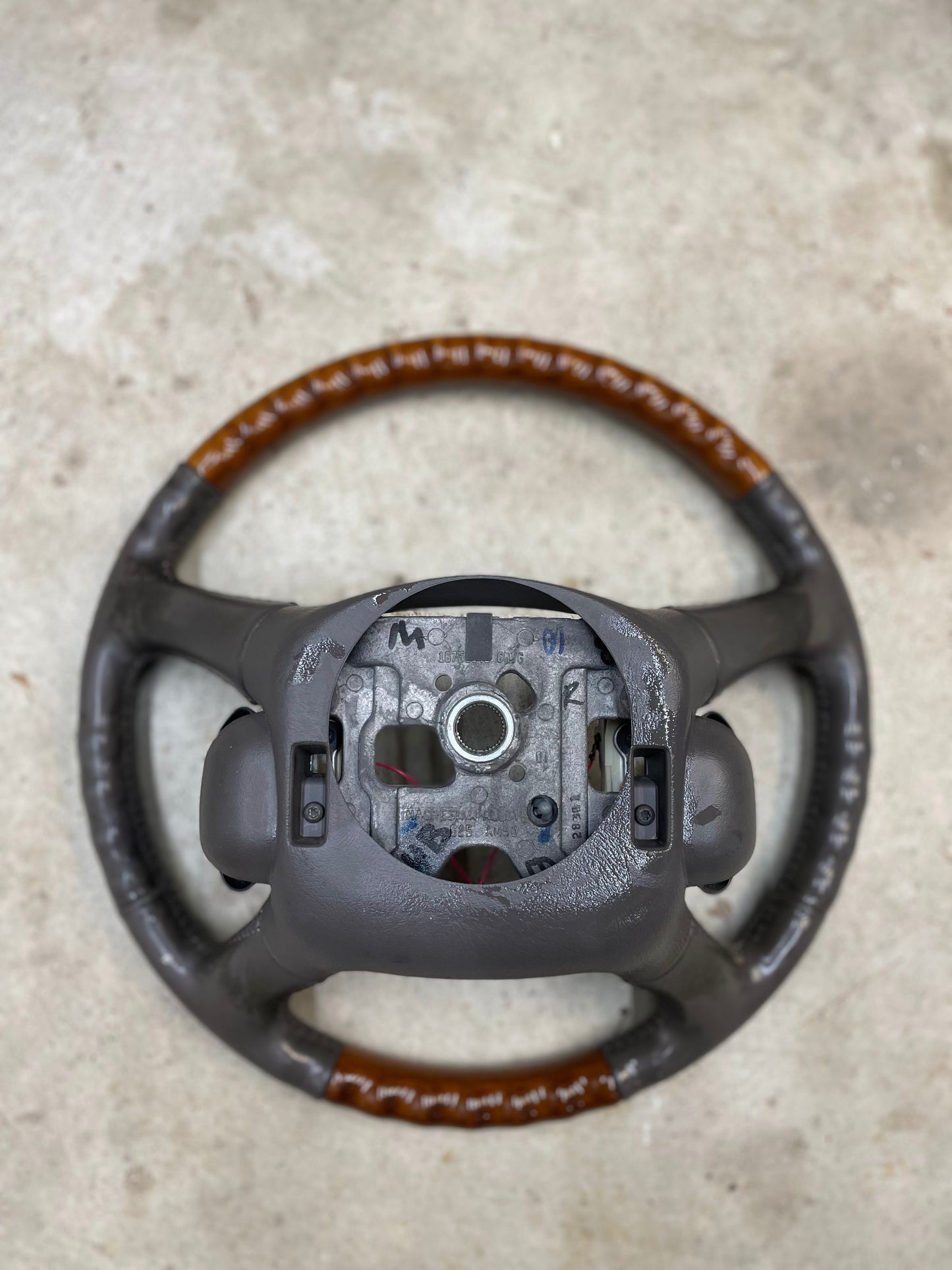 A worn General Motors OEM Leather w/Wood Steering Wheel with radio controls from an Escalade (fits 1995-2005 GM Trucks) is partially disassembled, with exposed metal and missing center, faded leather, and lies on a concrete surface.