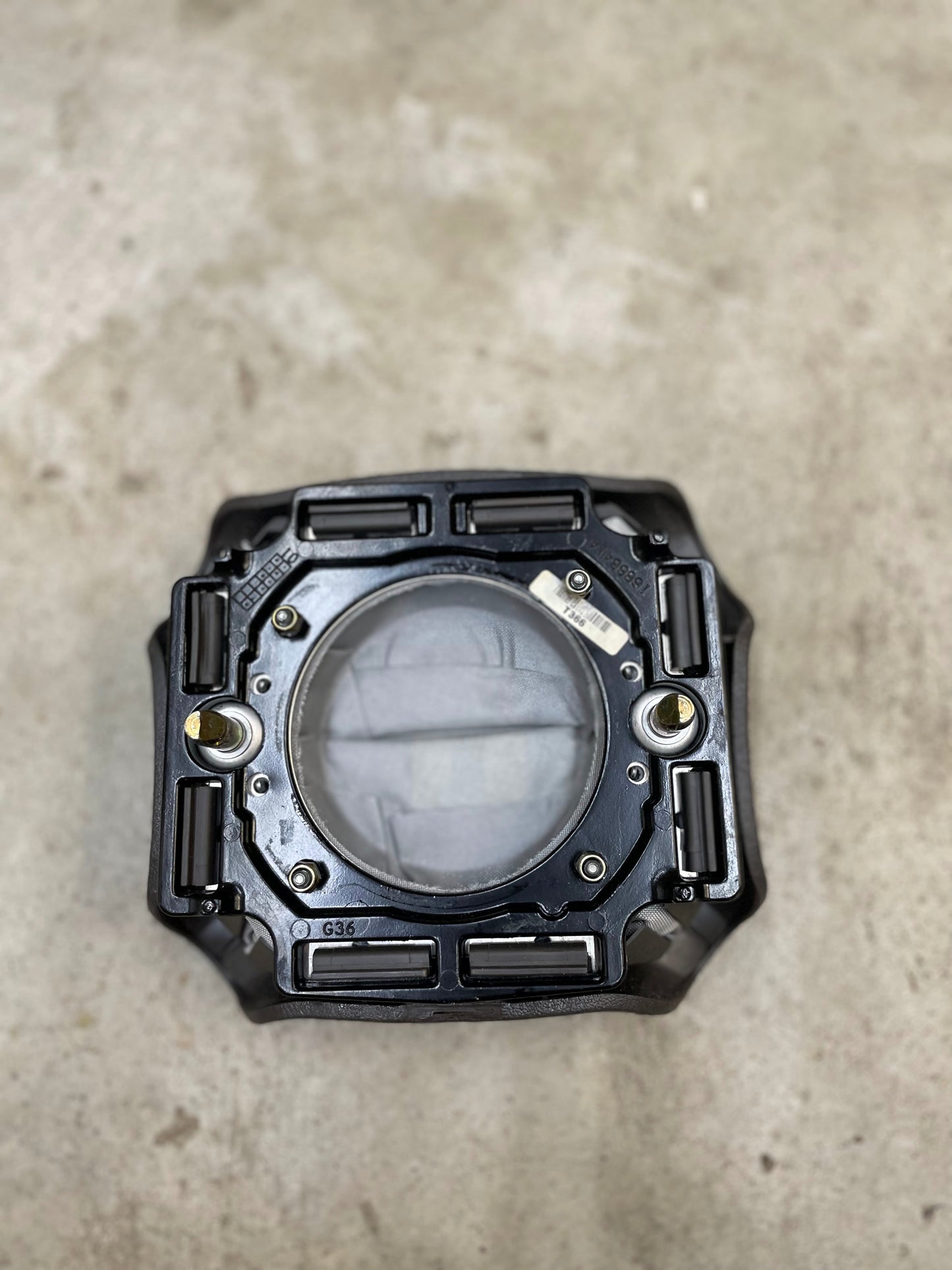 A black, rectangular mechanical part with a circular window, likely from the General Motors KIT Leather w/Wood Steering Wheel & Radio Controls for 1995-2005 GM trucks, sits on concrete and has bolts and grooves along its edges.