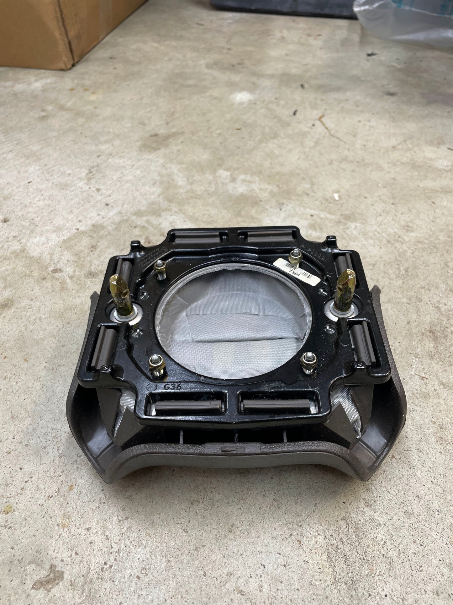 A General Motors KIT Leather w/Wood Steering Wheel with Radio Controls from an Escalade (fits 1995-2005 GM Trucks) sits on a concrete floor, featuring a circular center, black housing, and exposed bolts with boxes in the background.