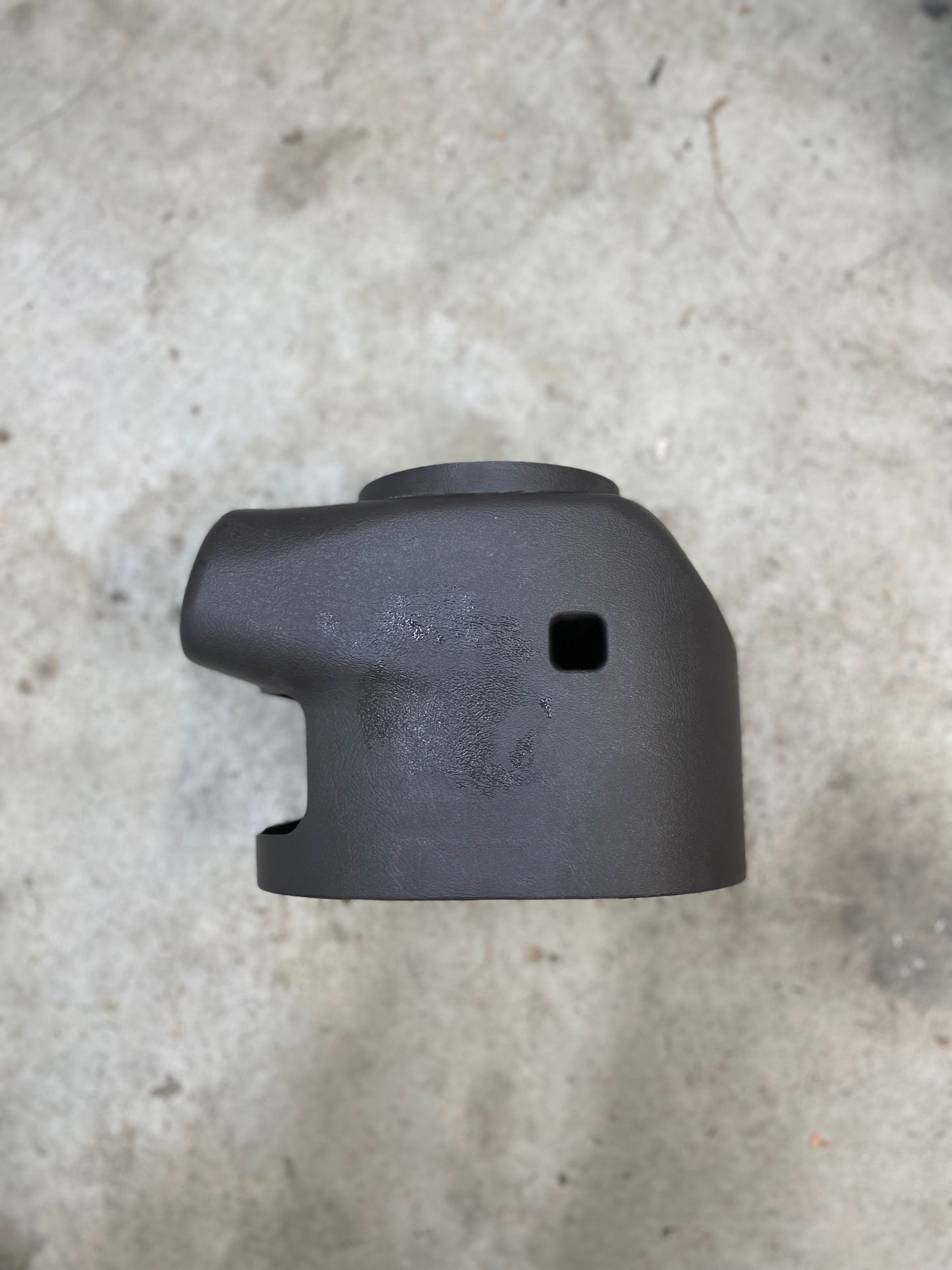 A matte black, cylindrical metal part with a rounded top and unique openings rests on a light concrete surface—designed for the KIT Leather w/Wood Steering Wheel & Radio Controls from Escalade (1995–2005 GM Trucks) by General Motors.