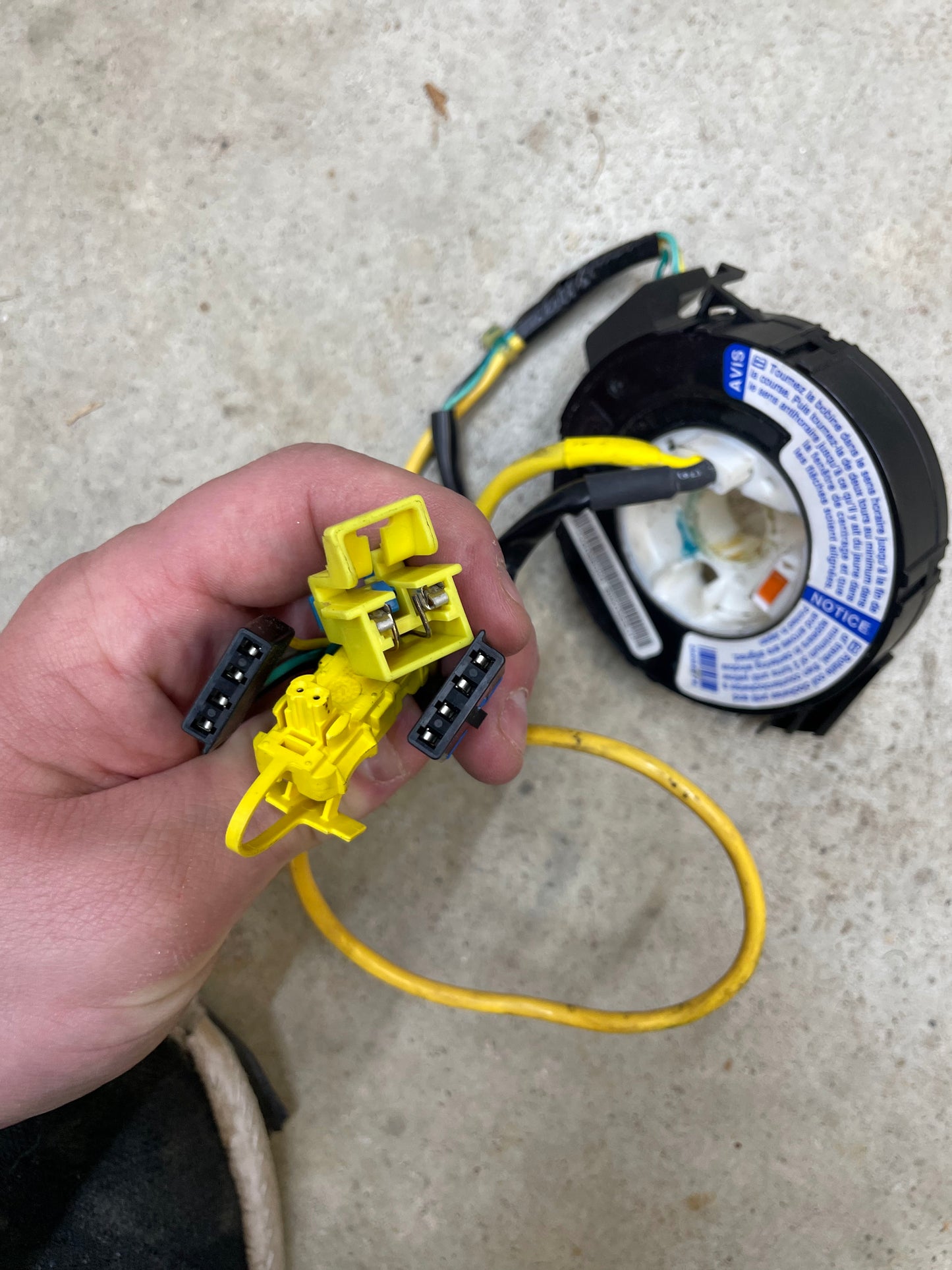 A hand holds yellow and black wiring connectors beside a circular car part with warning labels and a yellow cable, all on a light surface next to a General Motors KIT Leather w/Wood Steering Wheel & Radio Controls from Escalade for 1995-2005 GM Trucks.