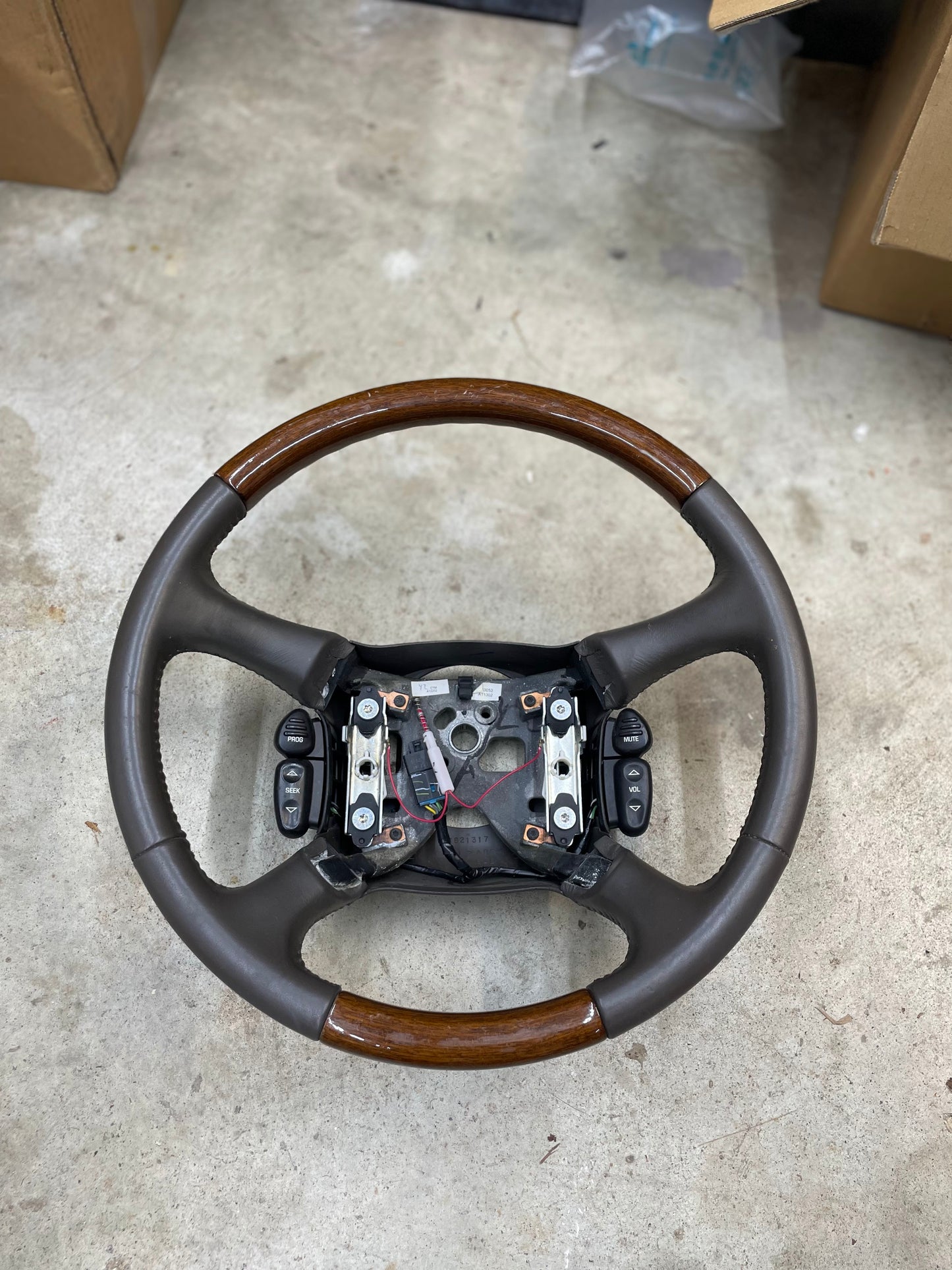 A General Motors KIT Leather w/Wood Steering Wheel with radio controls from an Escalade, designed for 1995–2005 GM trucks, features brown wood and black leather sections and is shown detached with exposed wiring and mounting points.