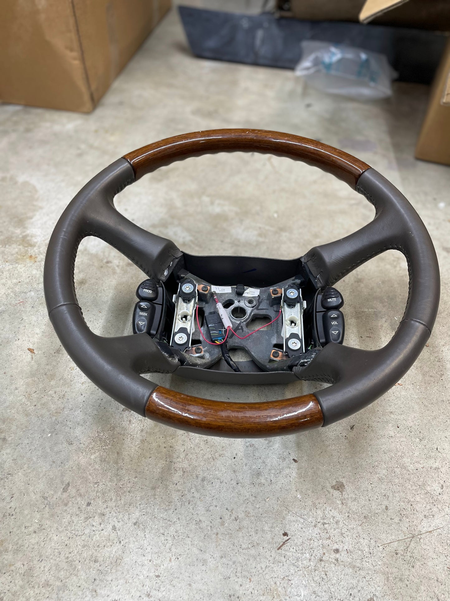 A General Motors KIT Leather w/Wood Steering Wheel with radio controls from an Escalade (fits 1995–2005 GM Trucks) rests on a concrete floor, with its center and airbag removed, wiring and mounting parts visible. Cardboard boxes are in the background.