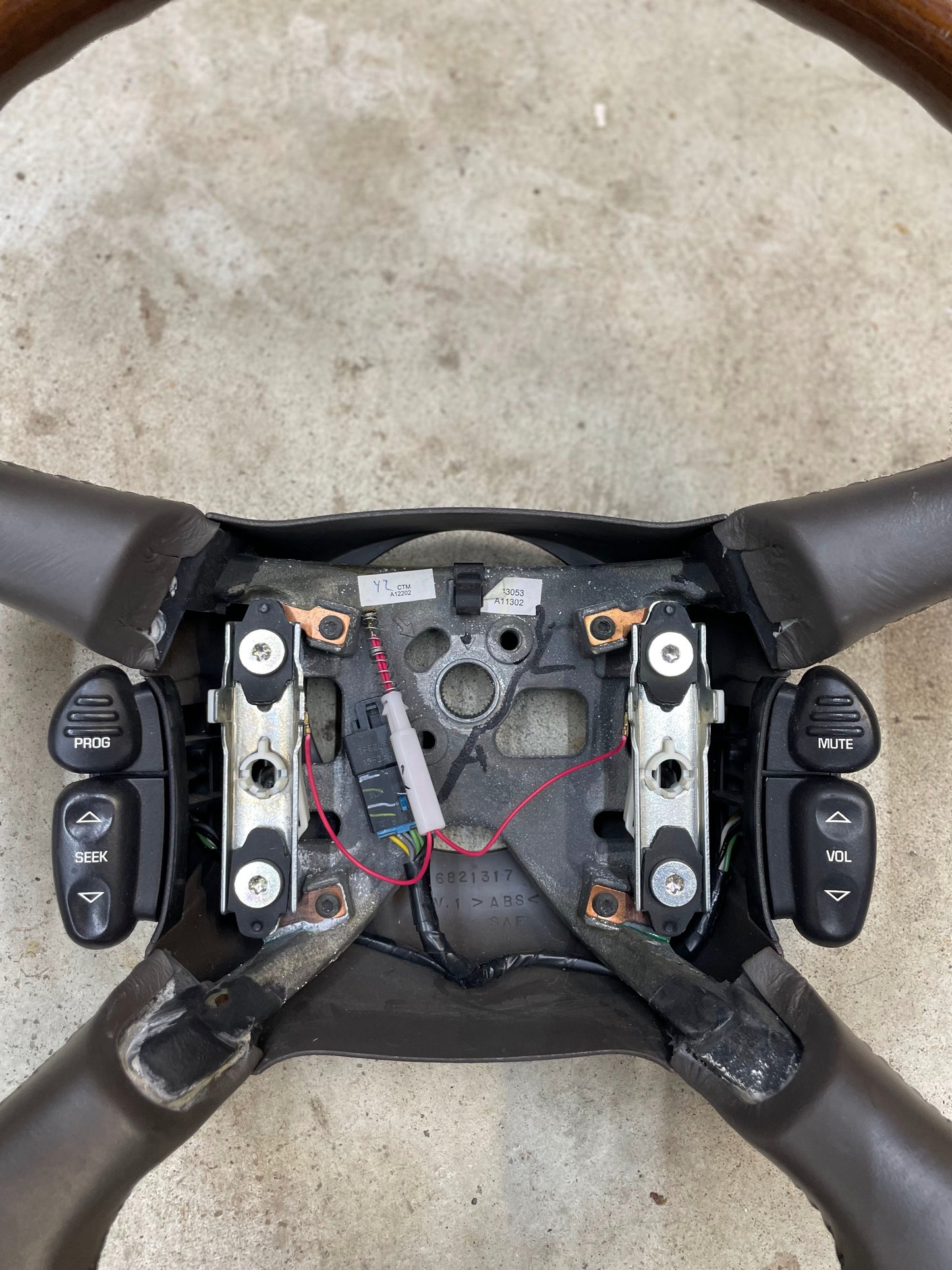 A General Motors KIT Leather w/Wood Steering Wheel & Radio Controls from Escalade (fits 1995-2005 GM Trucks) is shown disassembled, exposing wiring, metal parts, and black PROG/SEEK/VOL/MUTE buttons on a concrete surface.