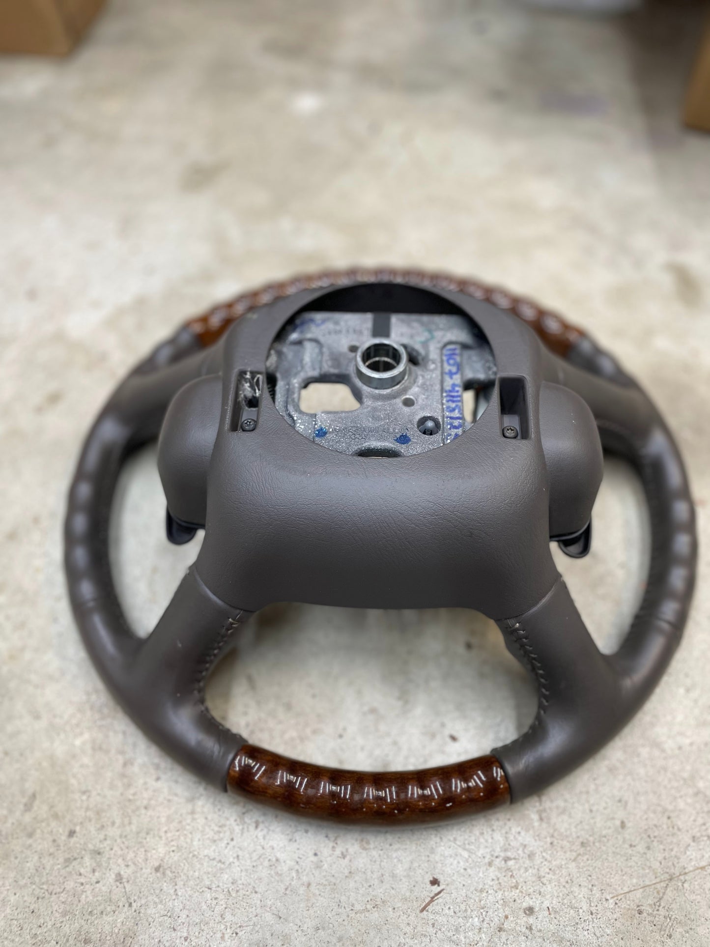 A General Motors KIT Leather w/Wood Steering Wheel & Radio Controls from Escalade (fits 1995-2005 GM Trucks) is shown on a concrete floor with the center panel and airbag removed, exposing the metal mounting area.