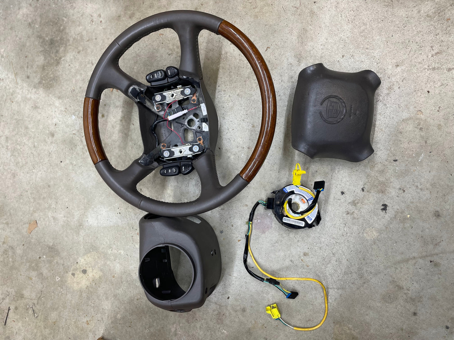A disassembled General Motors KIT Leather w/Wood Steering Wheel with radio controls from an Escalade (fits 1995-2005 GM Trucks), plus wiring, horn, airbag components, column cover, and airbag module, is laid out on a concrete surface.
