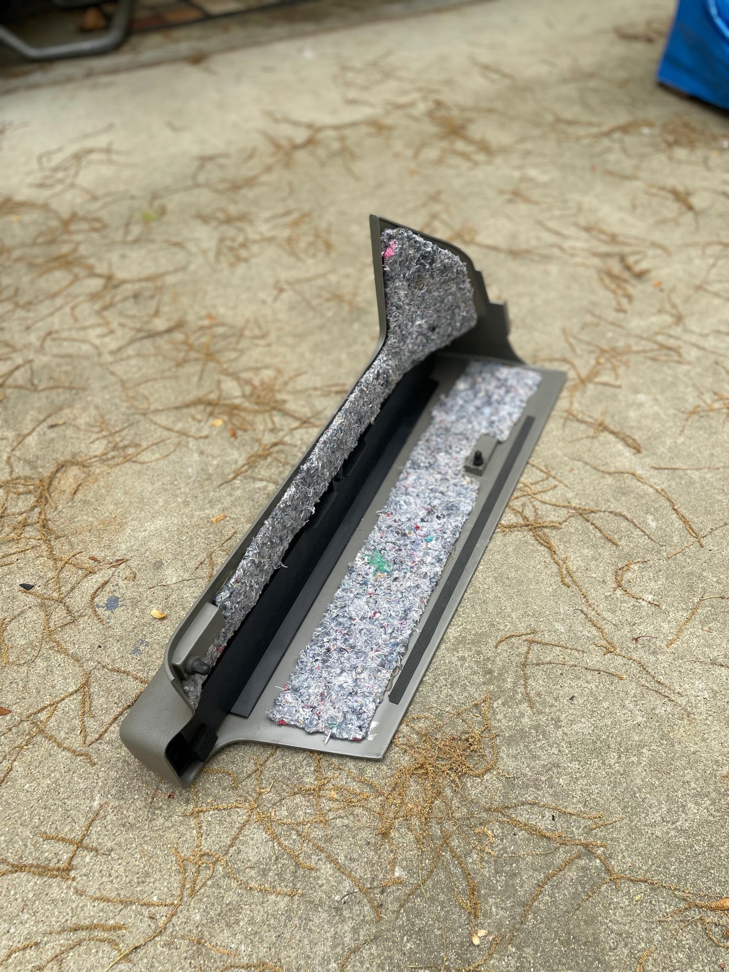 A NOS Retractable Rear Cargo Shade Jack Cover in gray for 2 Door 1995-2005 Chevy Blazer/GMC Jimmy by General Motors rests on a concrete surface covered with pine needles, lined with shredded multicolored insulation.