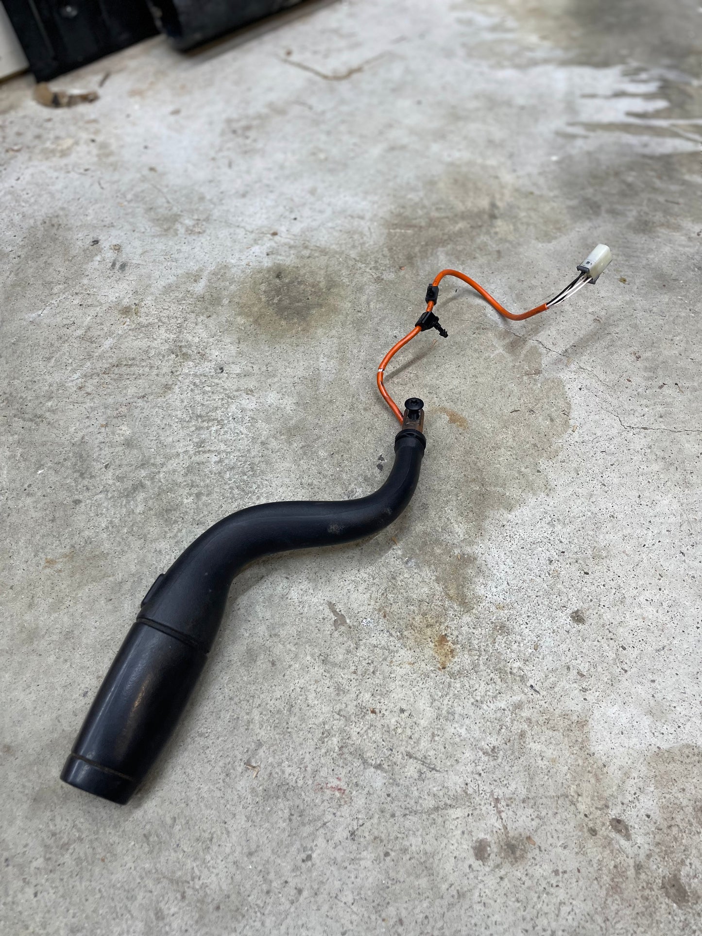 An OEM Column Shift Lever with Tow/Haul & Tapshift for 2006-2007 GM Duramax LBZ by General Motors, featuring a black curved pipe, orange and black wires, and a white connector, lies on a stained concrete floor.