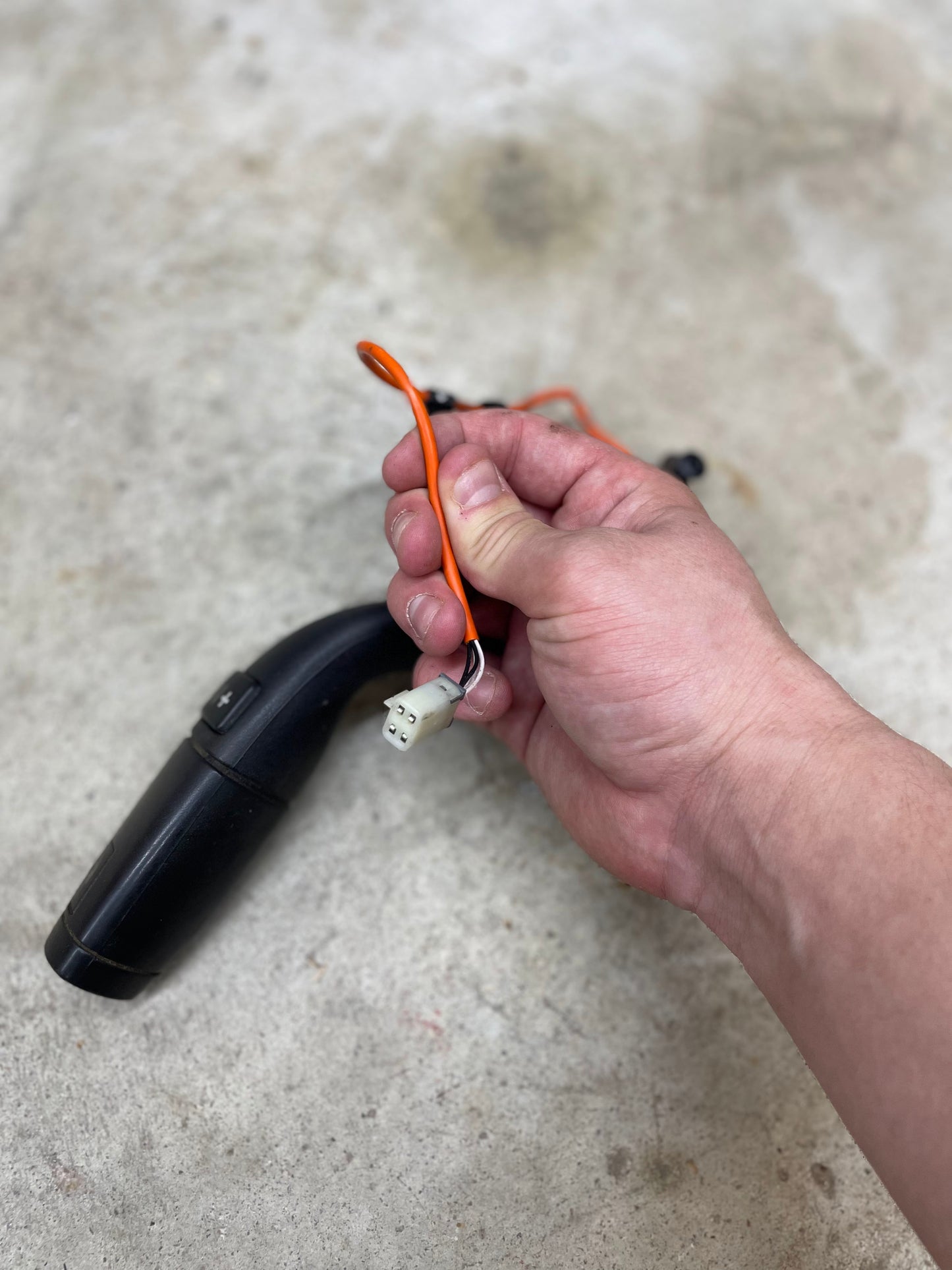 A hand holds an orange wire with a white connector attached to a General Motors OEM Column Shift Lever with Tow/Haul & Tapshift for 2006-2007 GM Duramax LBZ, set against a light, slightly textured background.