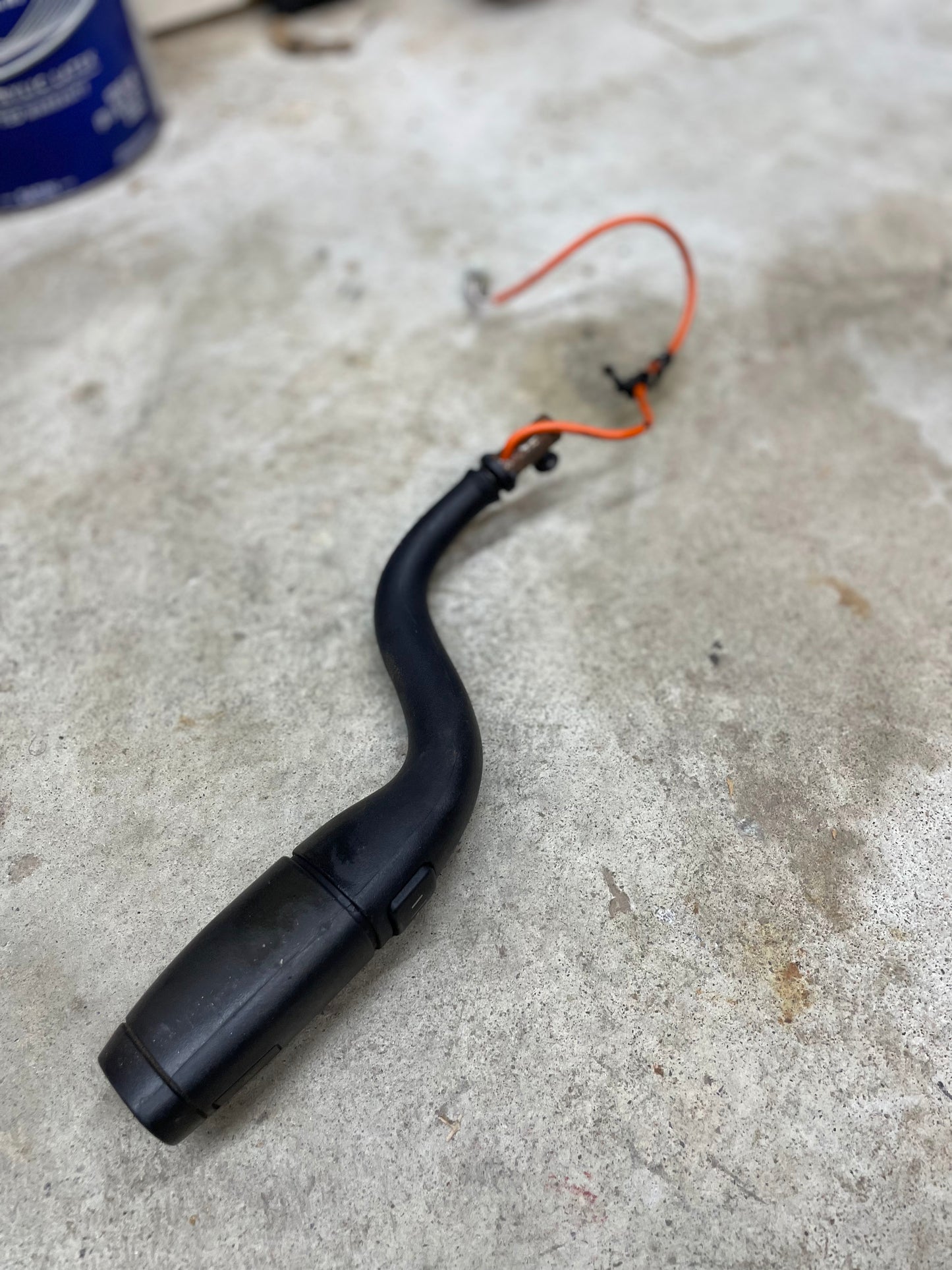 A General Motors OEM Column Shift Lever with Tow/Haul & Tapshift for 2006-2007 GM Duramax LBZ, featuring a black curved plastic design and attached orange-red wire, is shown lying on a concrete surface.