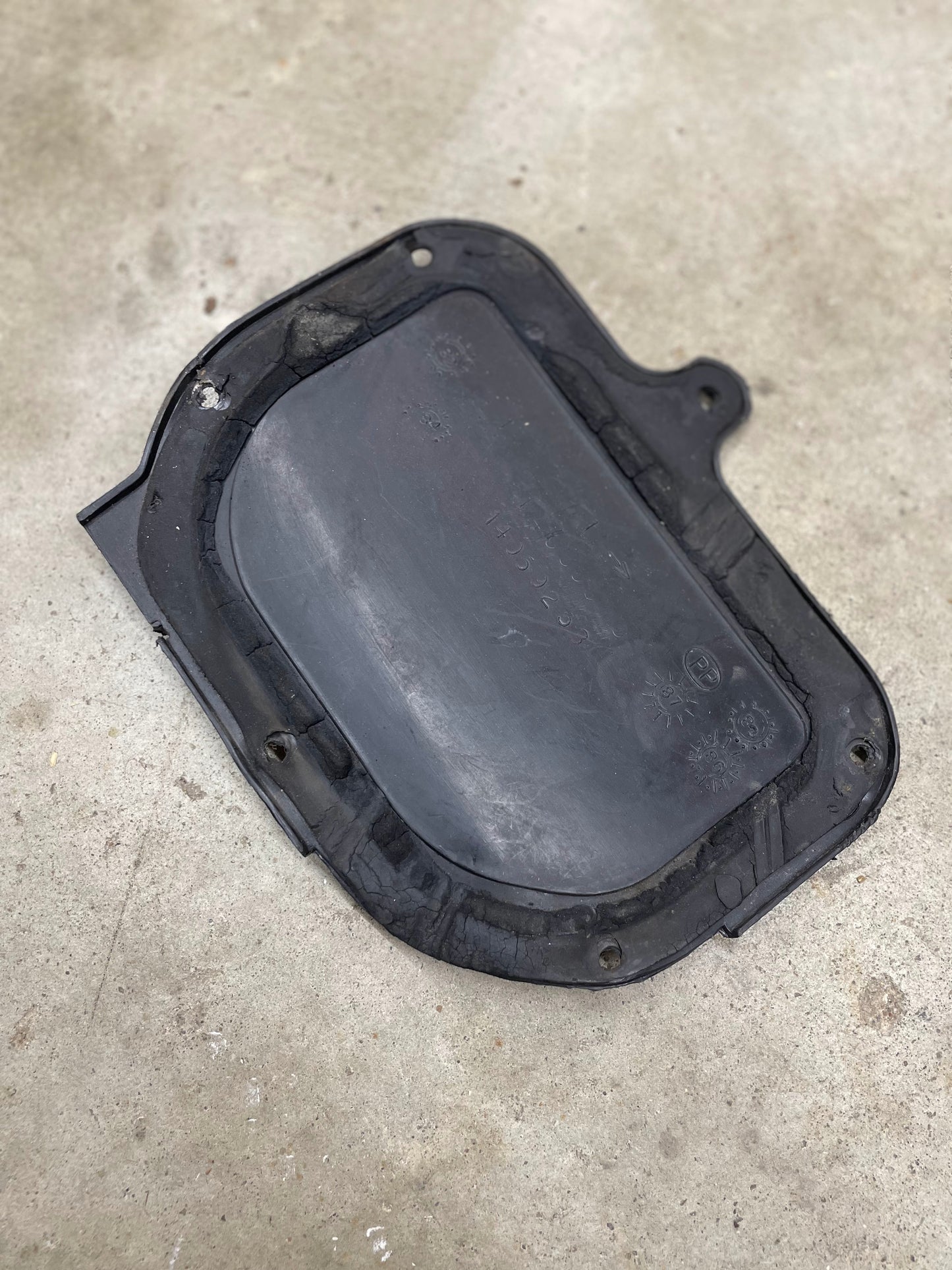 A worn OEM Fresh Air Block-Off Panel by General Motors for 1982-1993 Chevrolet S10, Blazer, and more—black rubber with rounded edges and a small tab—rests on a rough, light-colored concrete surface.
