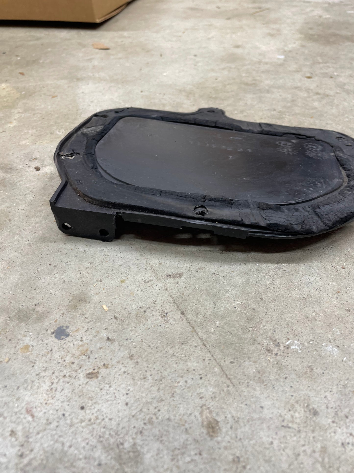 A black OEM Fresh Air Block-Off Panel by General Motors for 1982–1993 Chevrolet S10, Blazer, and more lies on a concrete floor. Slightly worn with screws and a raised border, it hints at prior use in automotive settings.