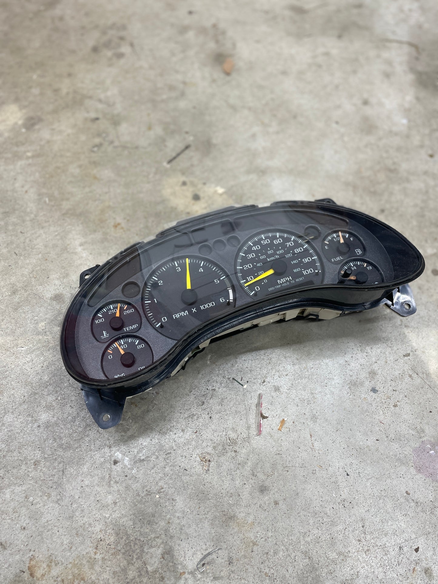 A General Motors OEM Tested Instrument Cluster with tachometer for 2001-2005 Chevy S10 or Blazer with console shifter sits on a concrete floor. The yellow speedometer needle points to about 30 mph.