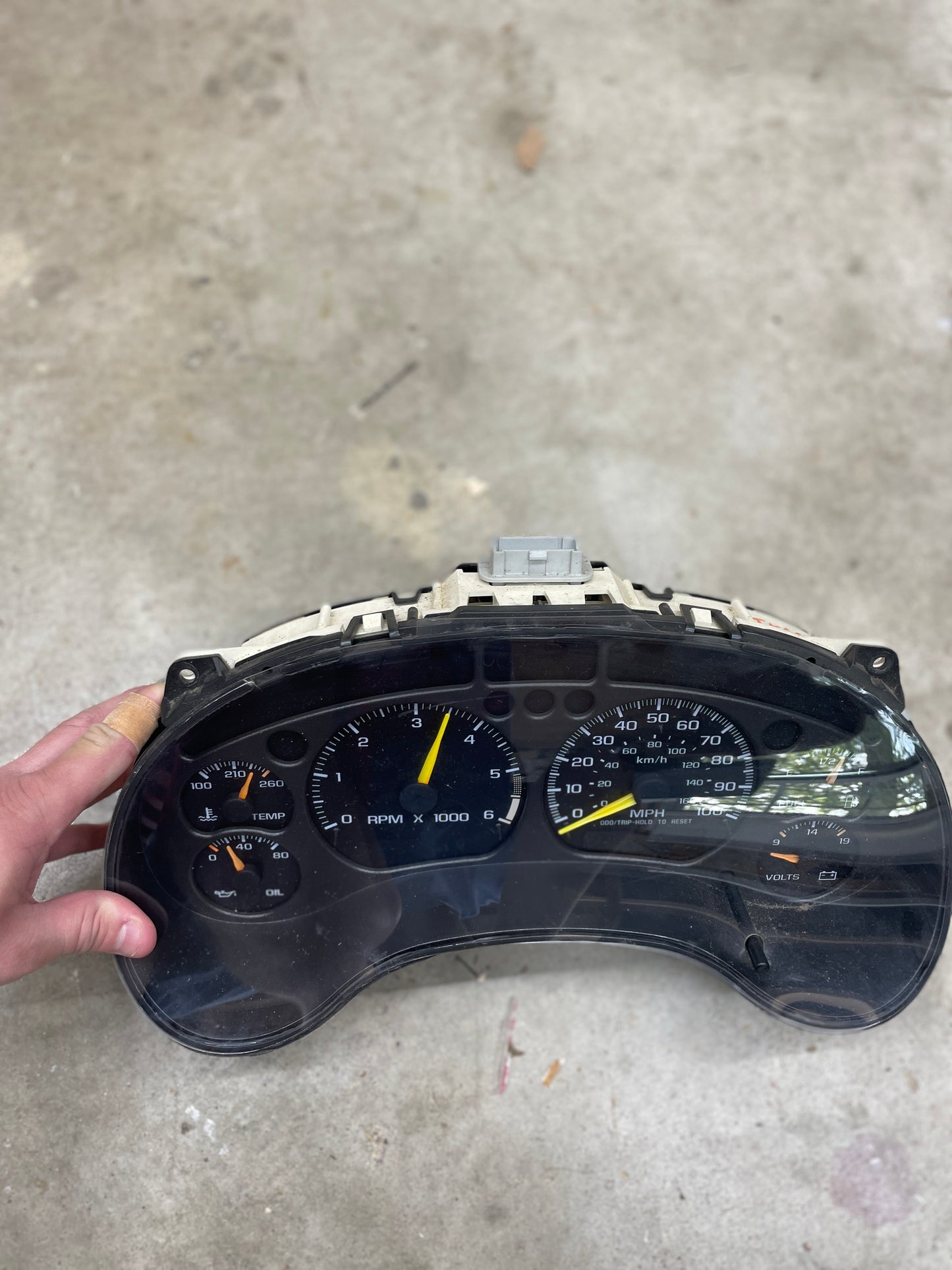 A hand holds a General Motors OEM Tested Instrument Cluster with Tachometer for 2001-2005 Chevy S10 or Blazer with Console Shifter, showing gauges for fuel, temp, RPM, MPH speed, oil pressure, and battery voltage against concrete.