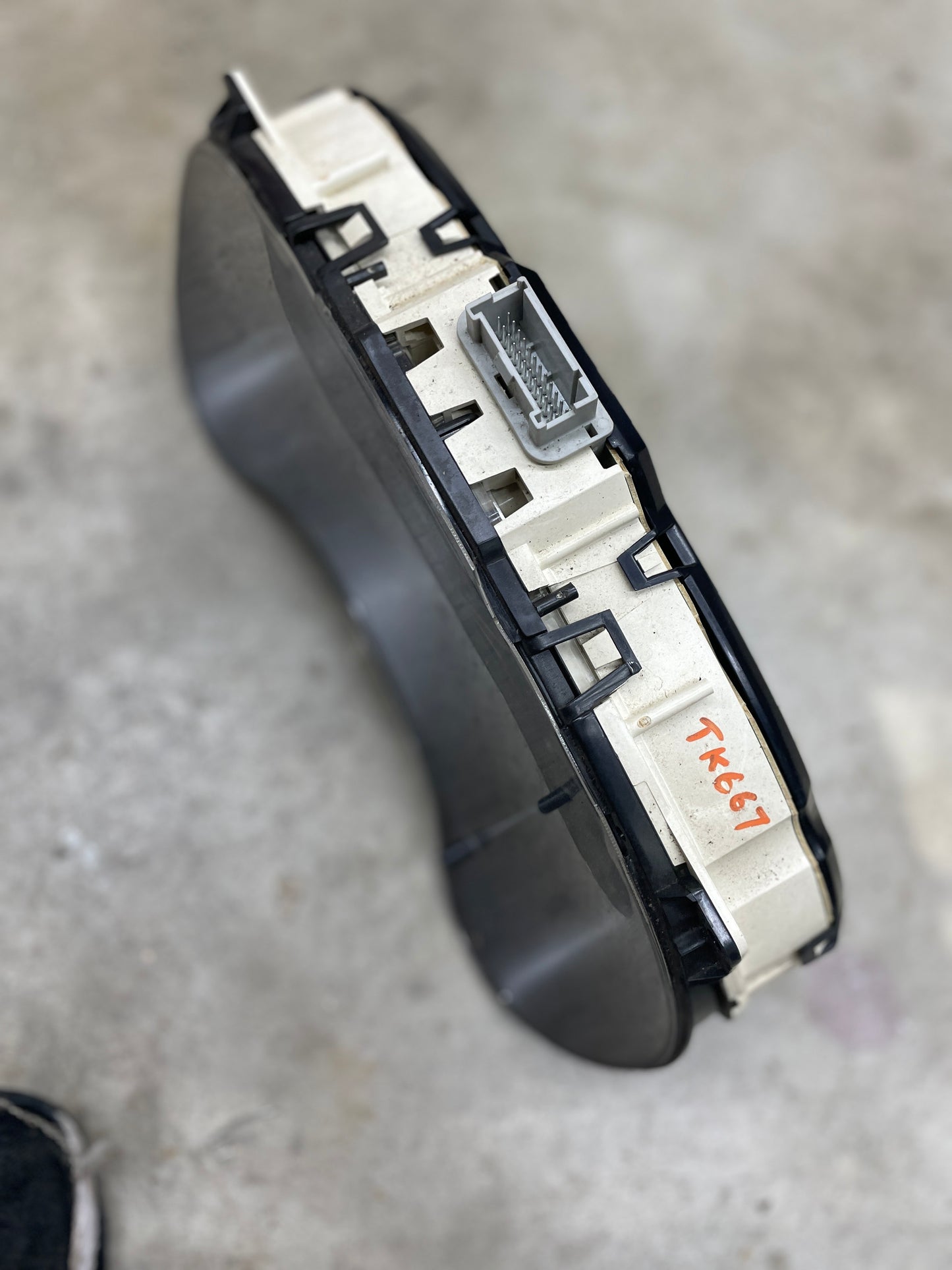 A close-up of a General Motors OEM Tested Instrument Cluster with Tachometer, code “1RK67” in orange on the white casing, for 2001-2005 Chevy S10 or Blazer with Console Shifter, shown on a gray surface with electrical connector.