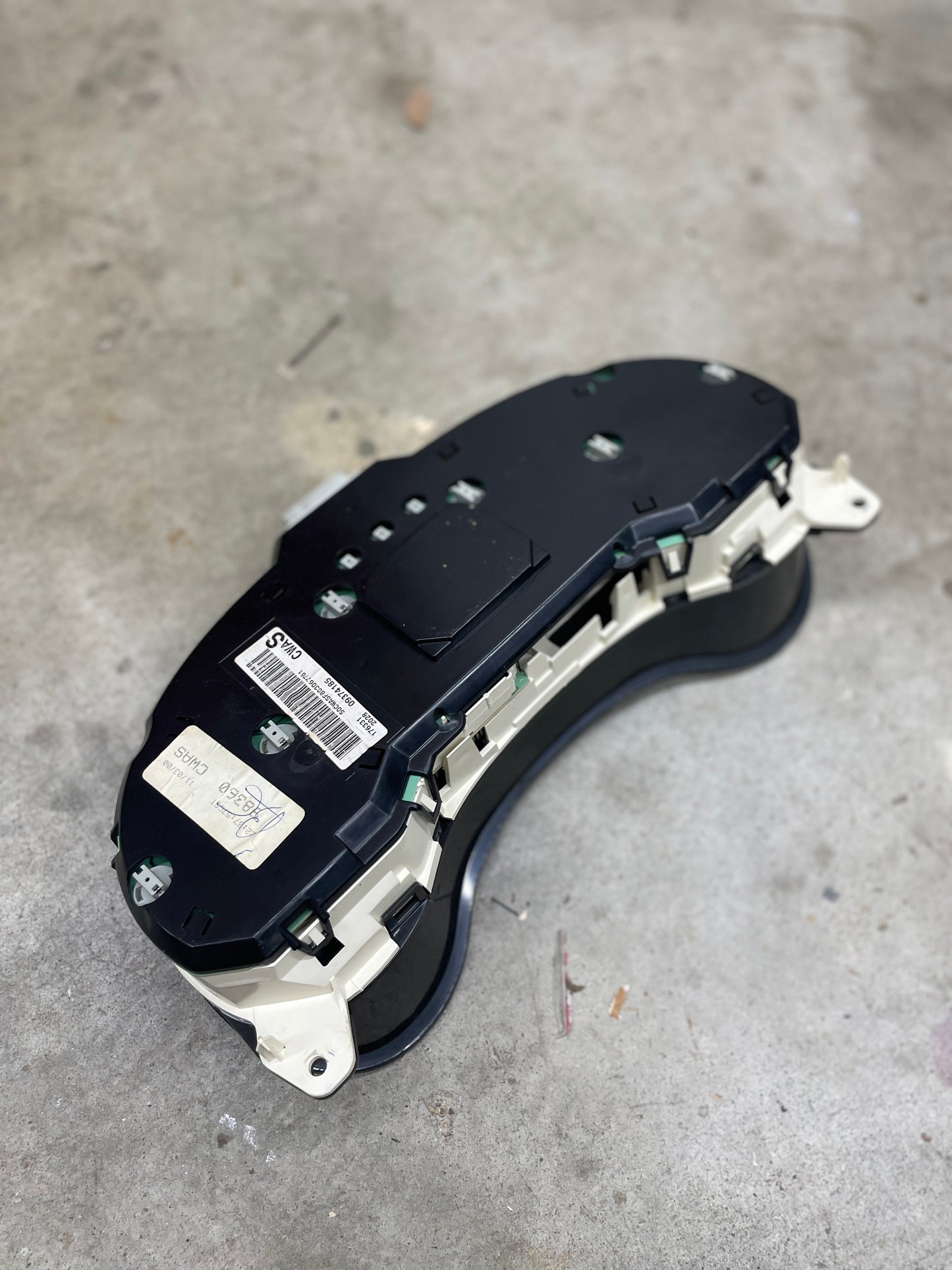 A General Motors OEM-tested instrument cluster with tachometer for 2001-2005 Chevy S10 or Blazer with console shifter is shown from the back, displaying its labeled black and white plastic casing on a concrete surface.