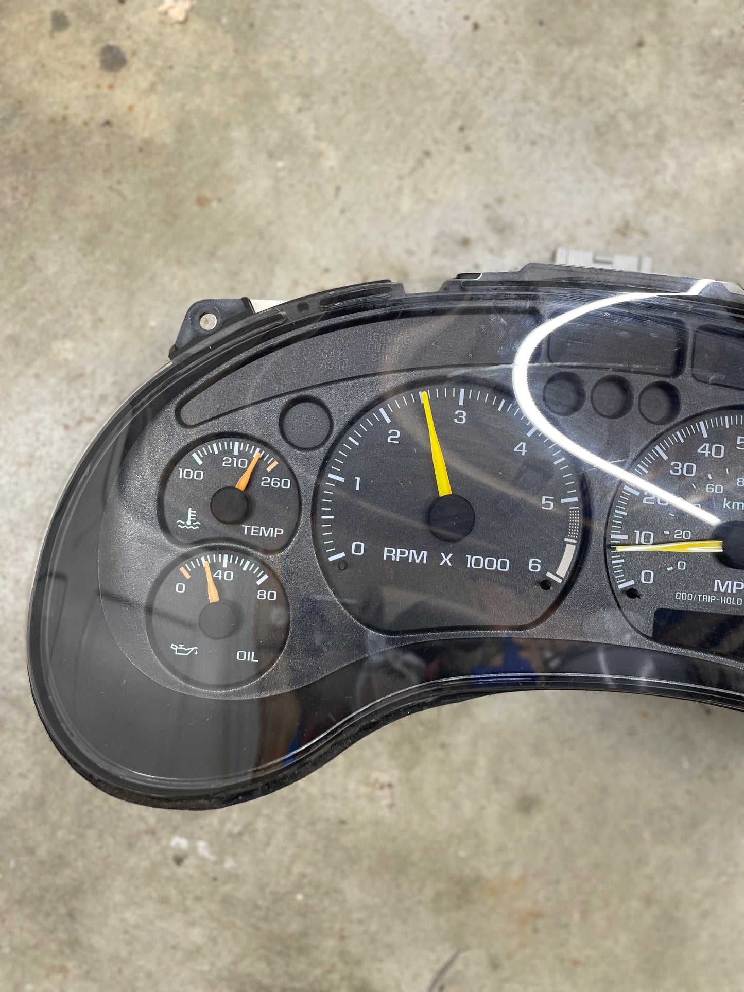 A close-up of a General Motors OEM Tested Instrument Cluster with Tachometer for 2001-2005 Chevy S10 or Blazer with Console Shifter shows oil, temp, RPM gauges (at ~2,800), and speedometer, with part of the dashboard above a concrete floor in the background.