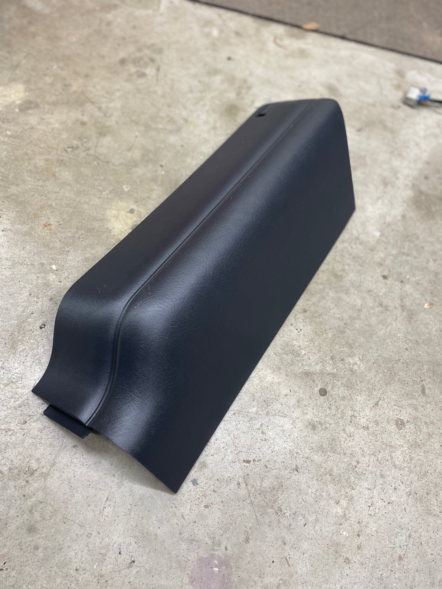 A dark gray, curved NOS Rear Compartment Jack Cover by General Motors for 4-door 1995-2004 Chevy Blazer and GMC Jimmy rests on a concrete floor in a workshop setting.