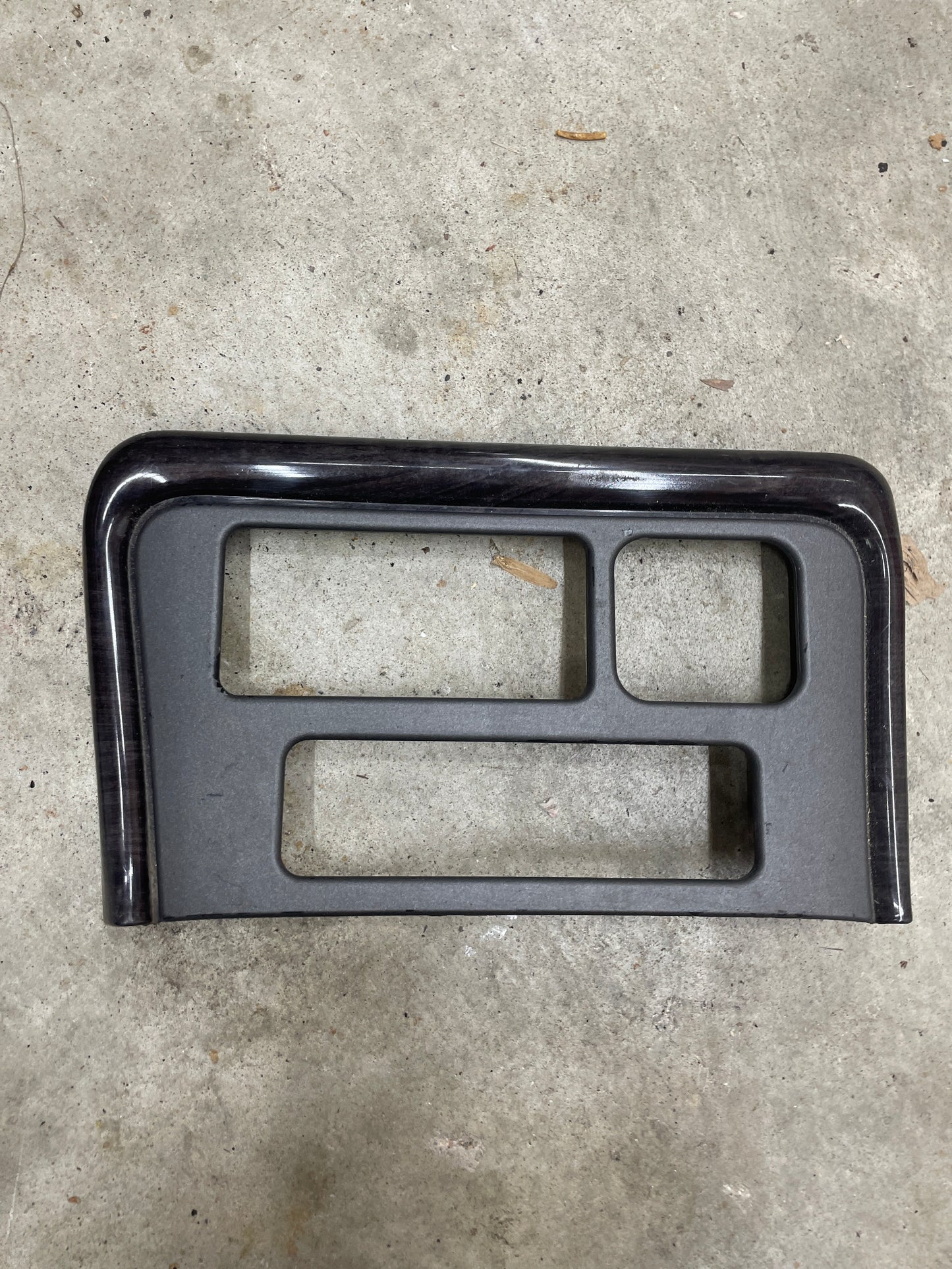 A General Motors OEM Center Console Front Bin Dark Woodgrain Trim for 2001-2002 GMC Sierra C3 or Yukon Denali is shown lying on a concrete floor.
