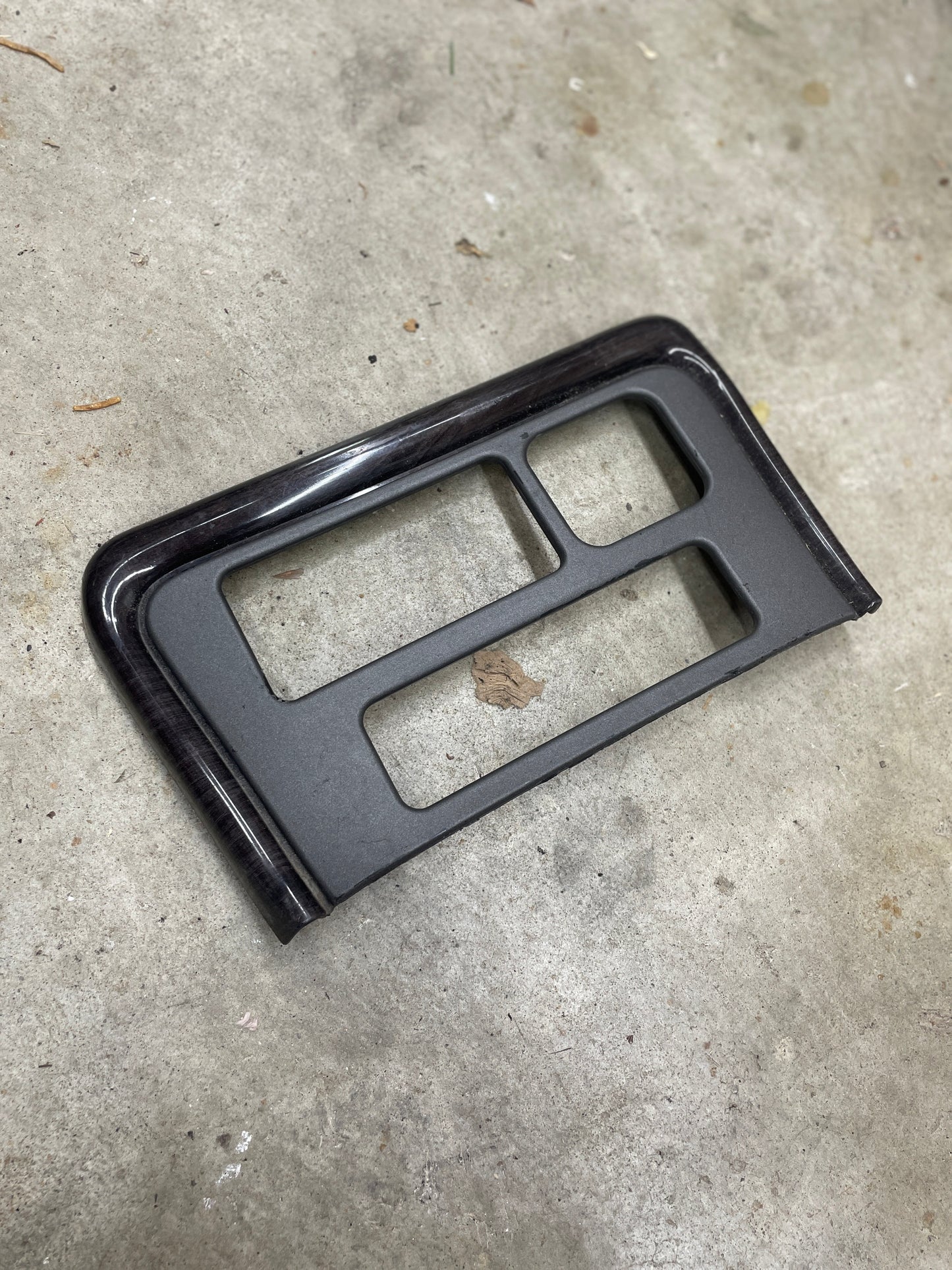 A General Motors OEM Center Console Front Bin Dark Woodgrain Trim for 2001-2002 GMC Sierra C3 or Yukon Denali, with four rectangular cutouts, rests on a gray concrete floor.