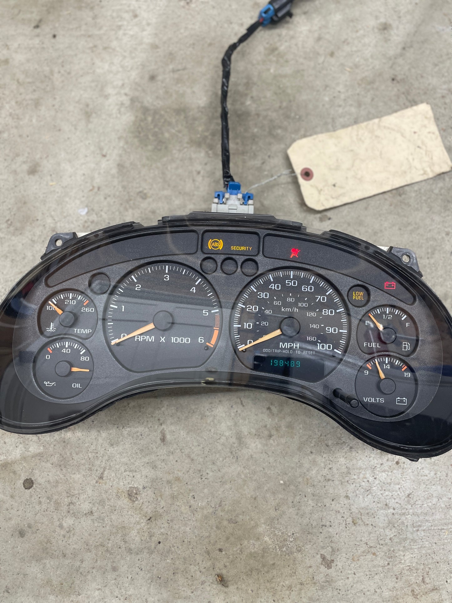 A General Motors OEM tested instrument cluster with tachometer for 2001-2005 Chevy S10 or Blazer (console shifter) sits on a concrete floor, tagged and equipped with analog gauges for RPM, speed, fuel, oil pressure, temp, and battery voltage.